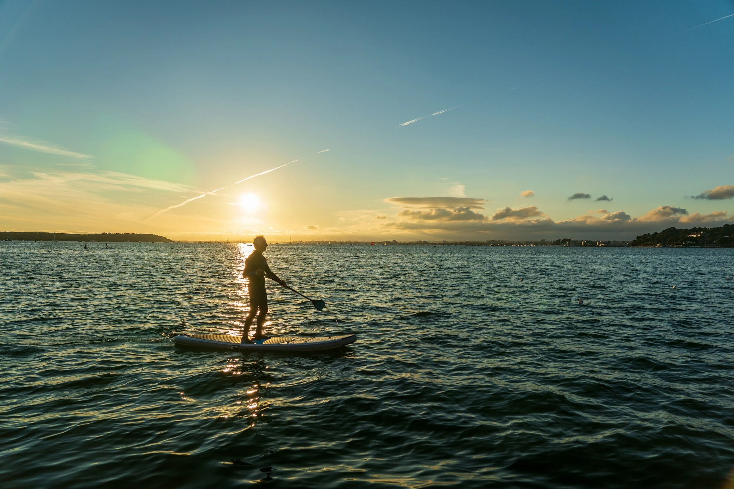 Person paddleboarding on a calm body of water during sunset, with a mostly clear sky and some clouds.