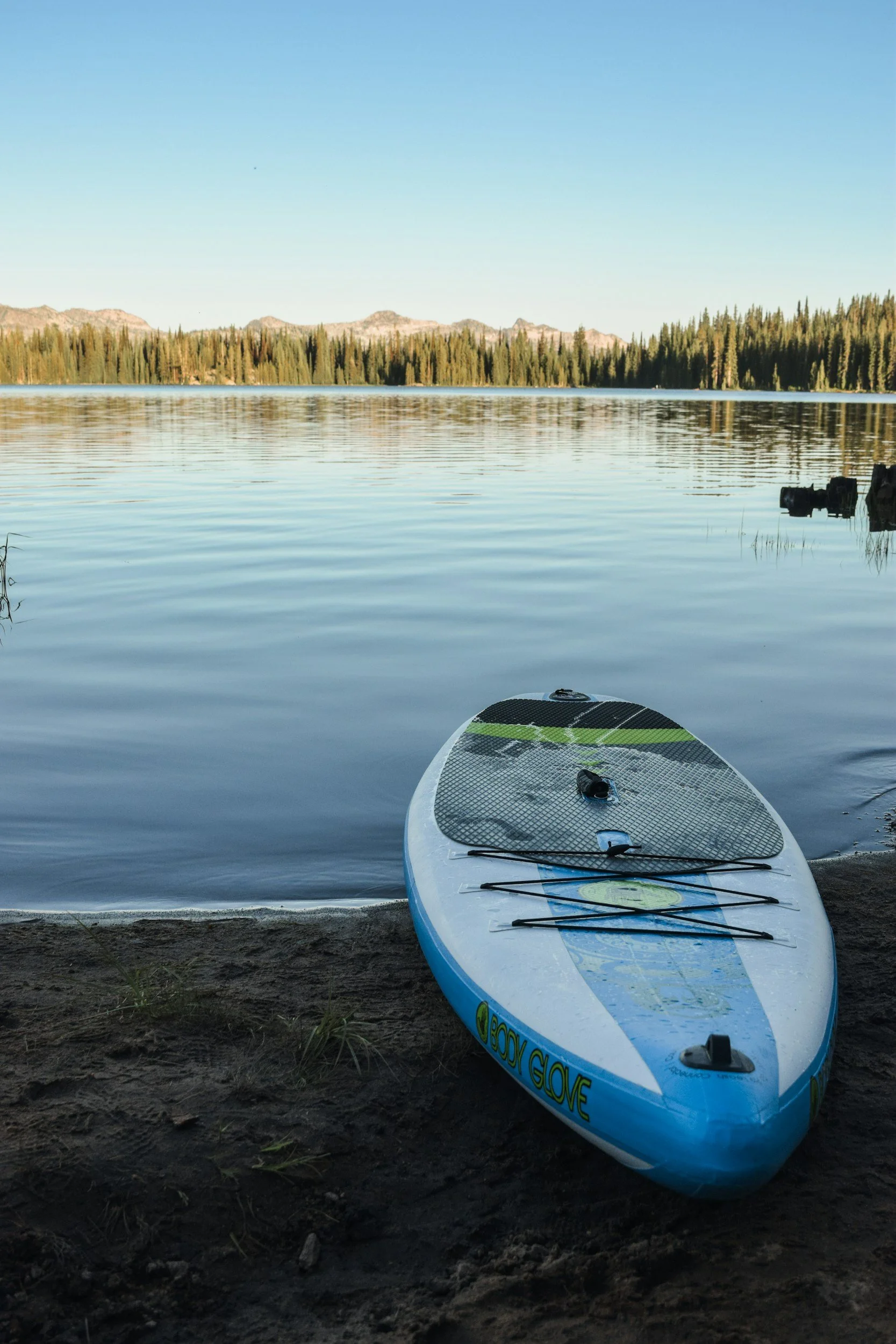 A paddleboard resting on a sandy shore beside a calm lake, with a forest and mountains in the background under a clear blue sky. Latta Nature Preserve, Mountain Island Lake & Lake Norman.
