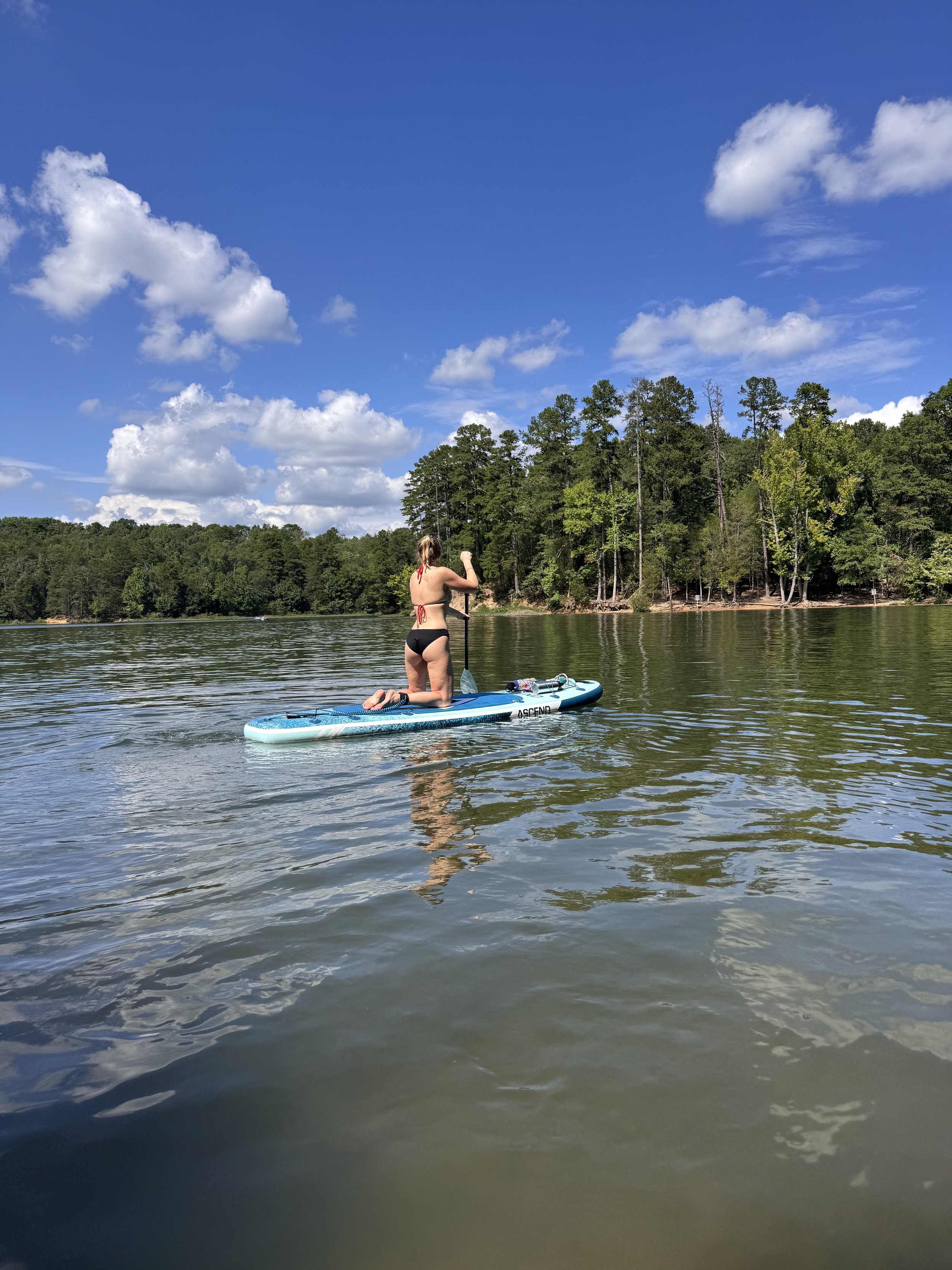 A woman in bikini kneeling on a paddleboard on a lake, paddling with a long-handled oar, with trees and blue sky with clouds in the background.