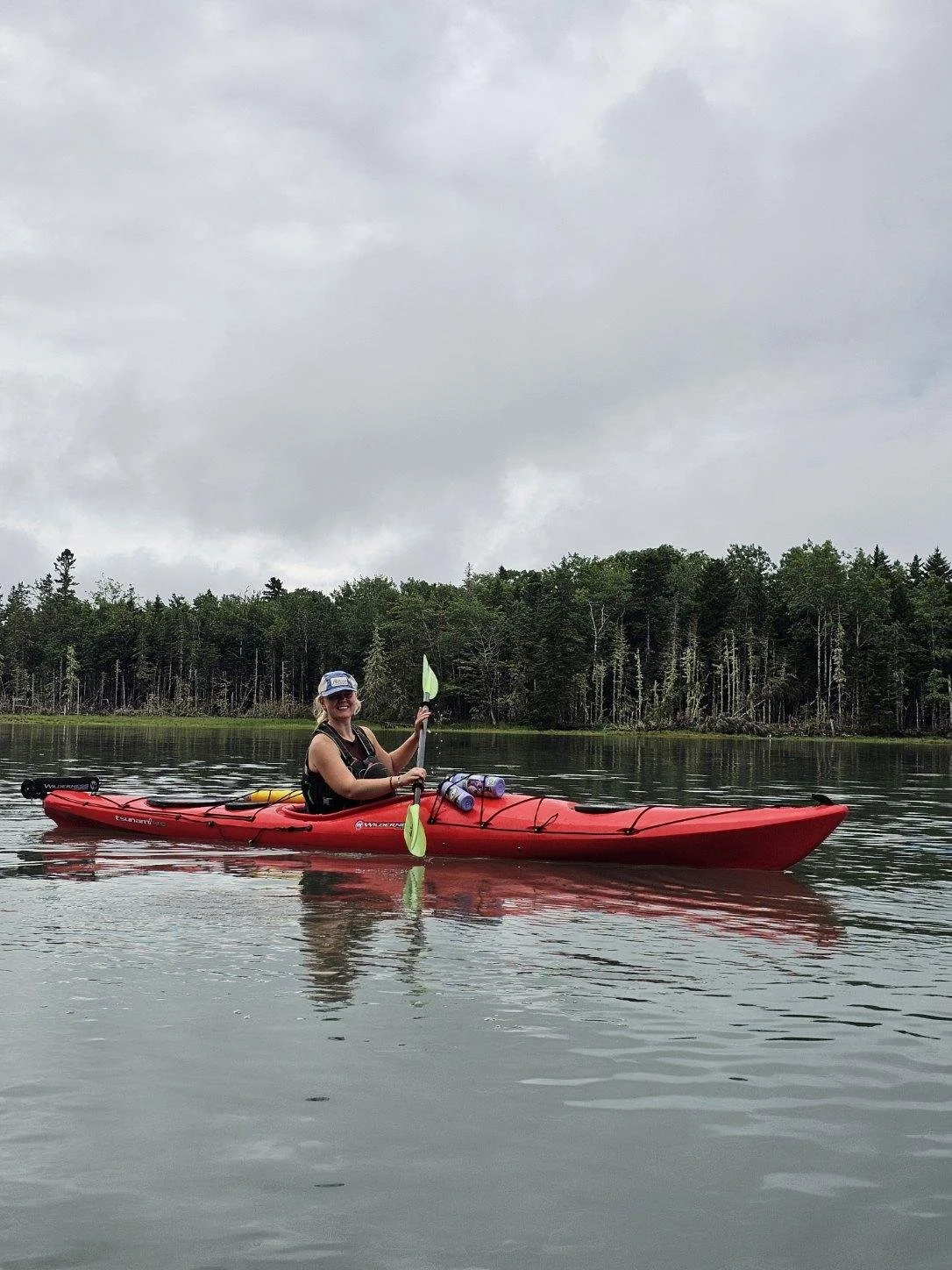 A woman in a kayak on a calm body of water holding a paddle, with a forested shoreline and cloudy sky in the background.