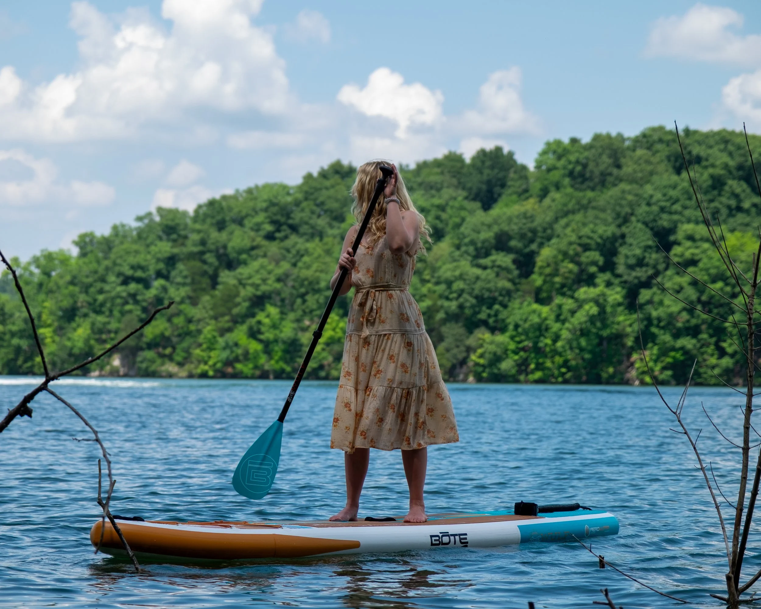 A woman standing on a paddleboard on a lake, holding a paddle, with a lush green forest and blue sky with clouds in the background.