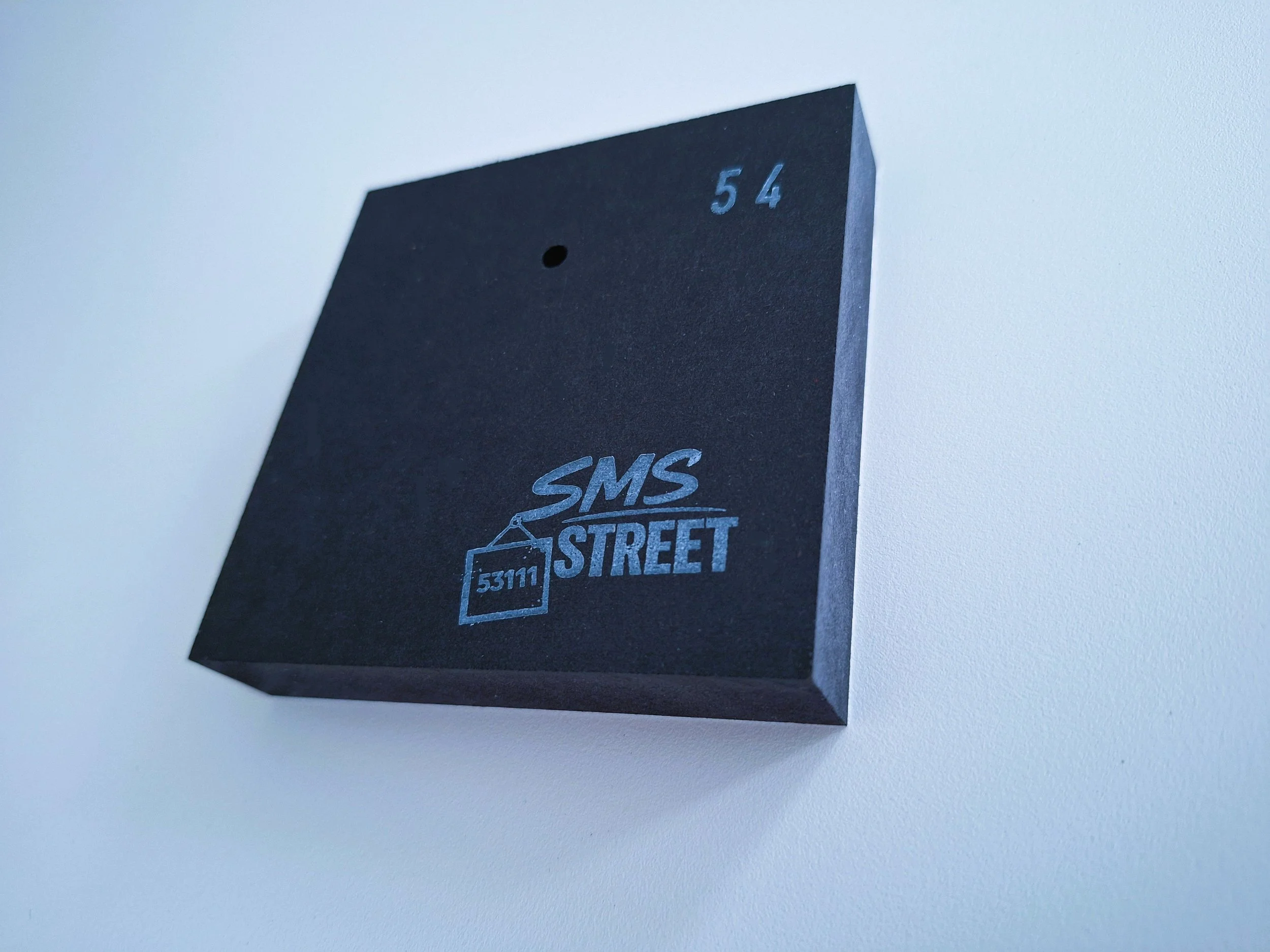 Schwarze Box mit blauer Aufschrift 'SMS STREET' und Hausnummer 54, auf weißer Wand montiert.