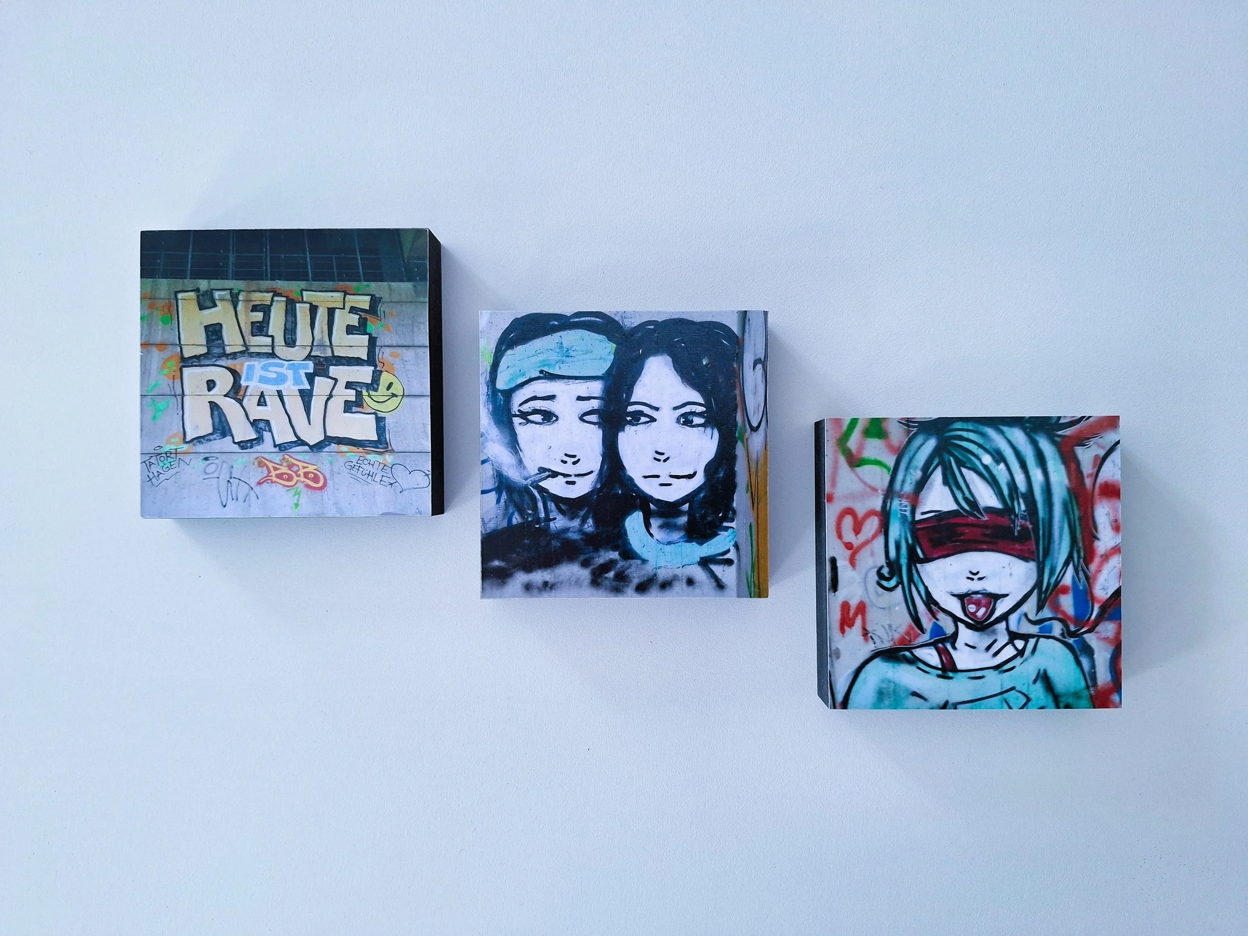 Drei Graffiti-Kunstwerke an einer weißen Wand: Das linke Bild zeigt den Schriftzug 'HEUTE IST RAVE' mit bunten Spritzern. Das mittlere Bild ist eine Schwarz-Weiß-Zeichnung zweier Frauen mit oben getragenen Kopfbedeckungen. Das rechte Bild zeigt eine 