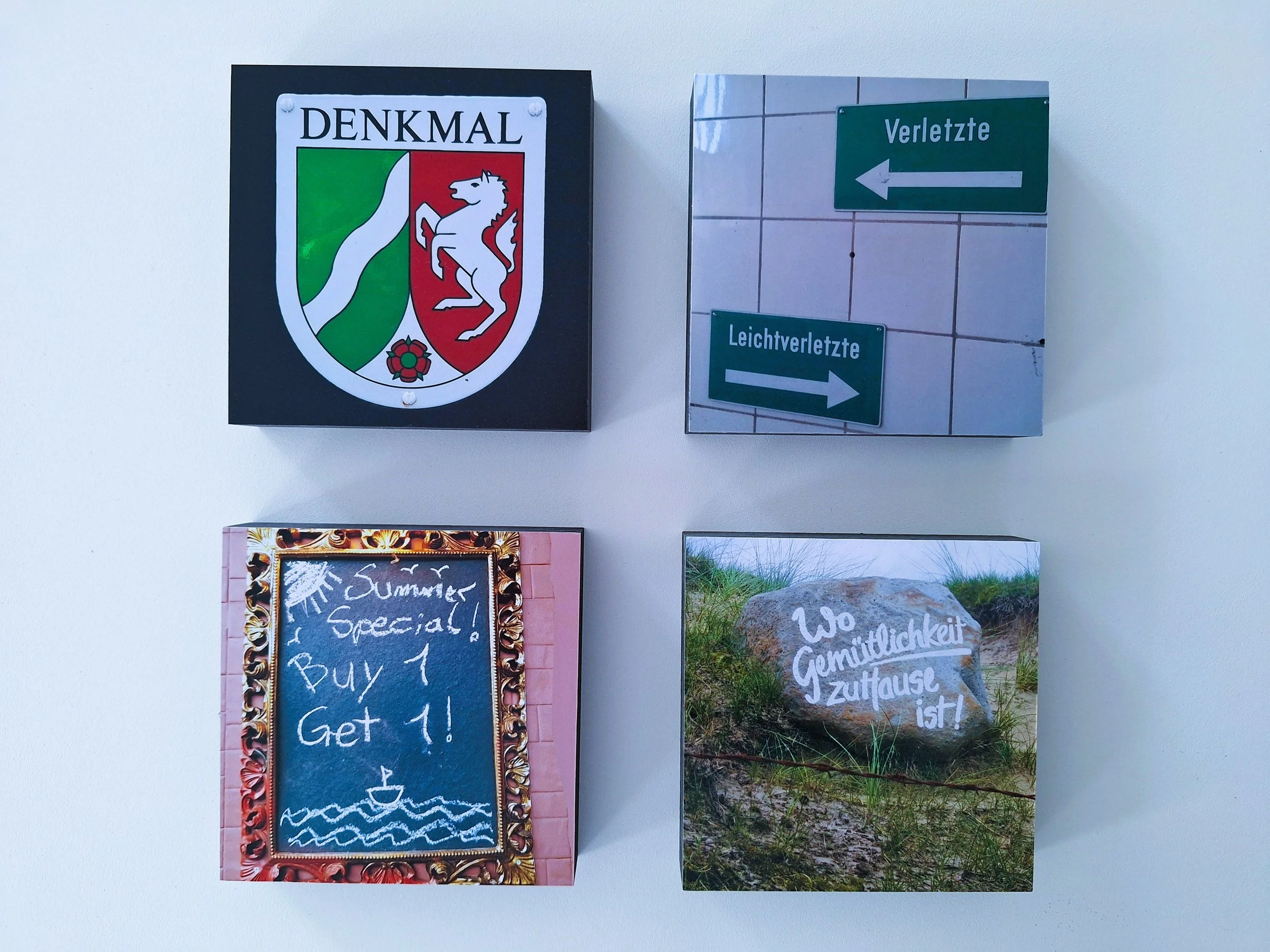 Vier quadratische Bilder auf weißer Wand. Das obere linke Bild zeigt ein Wappen mit der Aufschrift 'DENKMAL'. Das obere rechte Bild enthält grüne Richtungsbeschilder mit den Worten 'Verletzte' und 'Leichtverletzte'. Das untere linke Bild ist ein hand