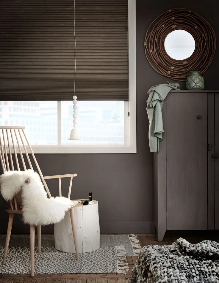 Interieur van een kamer met een groot raam, een houten stoel met een witte schapenvacht, een kleine witte bijzettafel met een nagellak en een lipstick, een kast met een groene vaas en een groene doek, decoratieve muurcirkel, donkergrijze muur en een bed met een zwarte en witte deken.