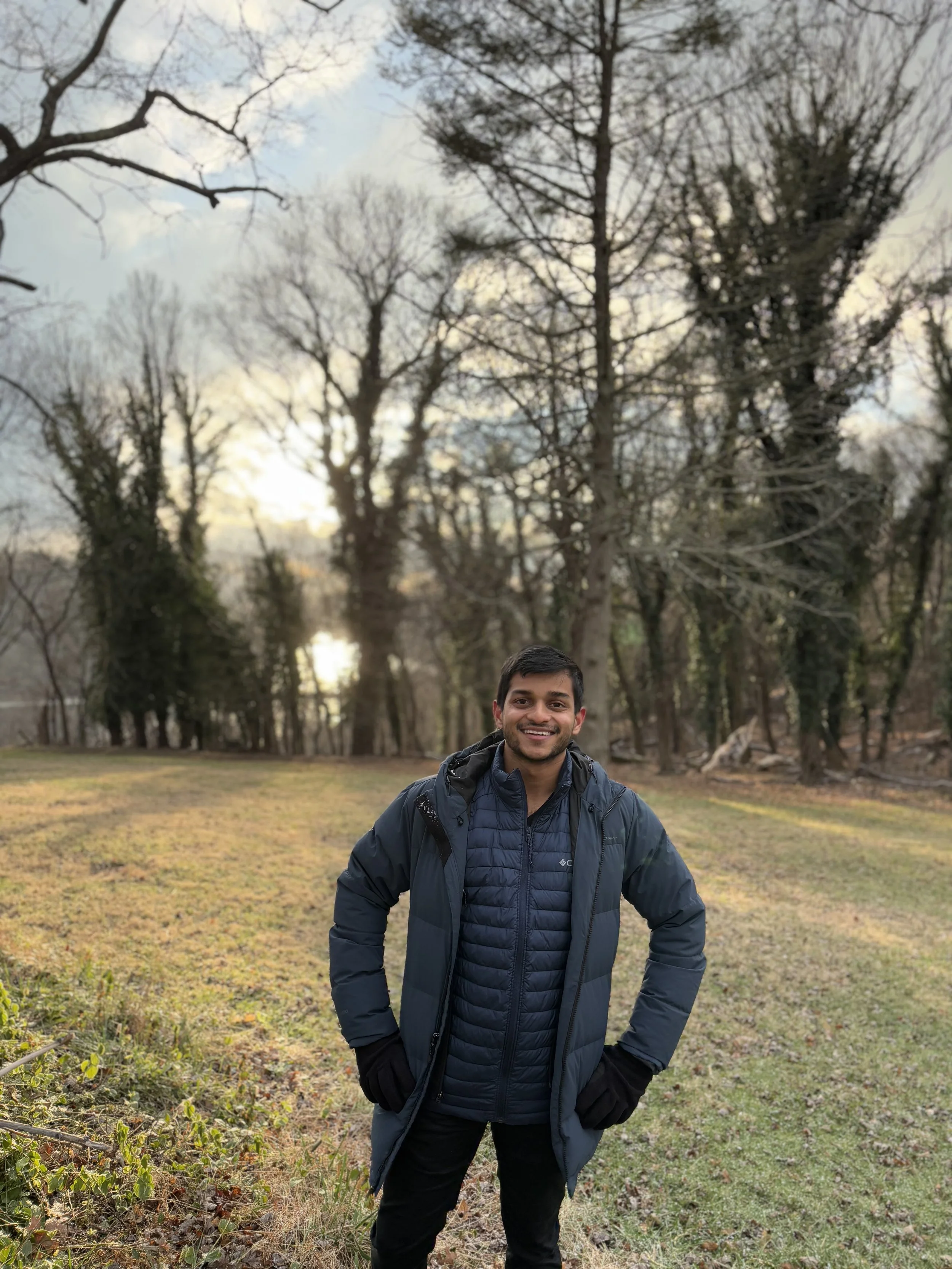 A smiling man wearing a dark jacket and gloves standing outdoors in a grassy area with leafless trees in the background during late fall or winter.