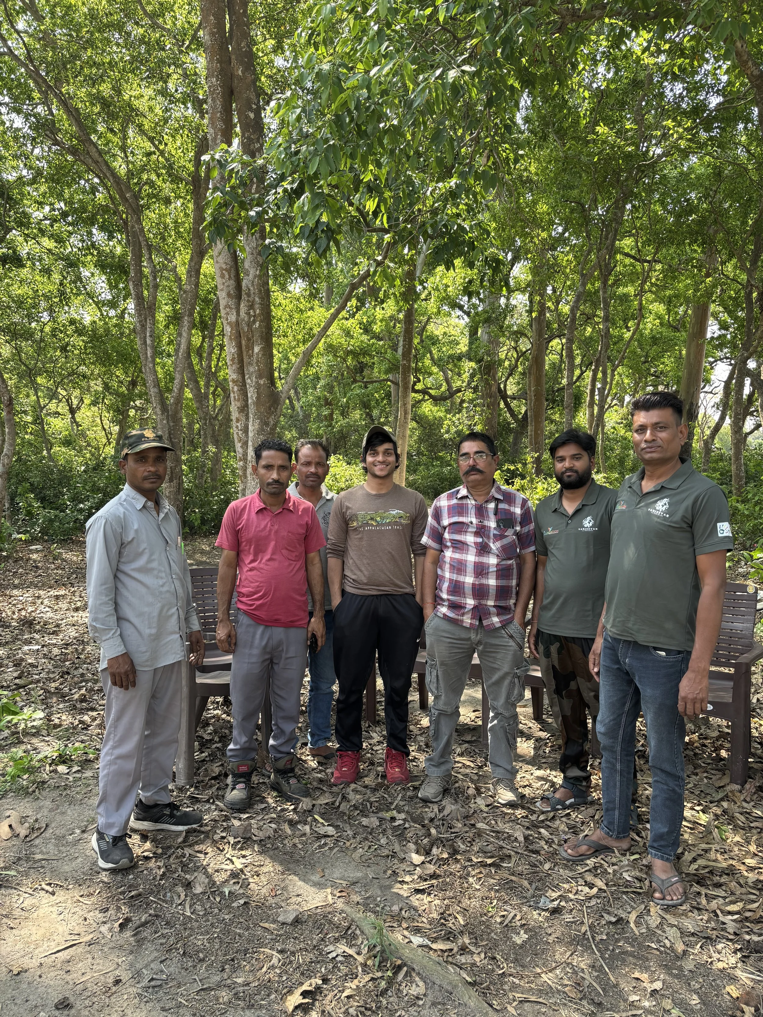 Group of seven men standing outdoors in a forested area with tall trees and green foliage, some sitting on benches behind them.