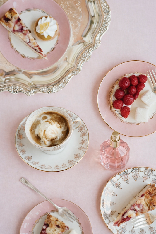 Elegant tea party setup with slices of berry cake on pink and floral plates, a cup of coffee with whipped cream, fresh raspberries on a cake, a small pink perfume bottle, and a silver mirror, all on a pink tablecloth.