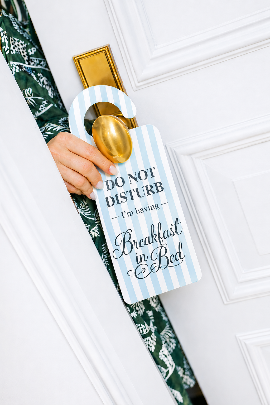 Close-up of a person holding a door hanger that says 'Do Not Disturb, I'm having Breakfast in Bed,' with a gold door handle and white paneled door in the background.