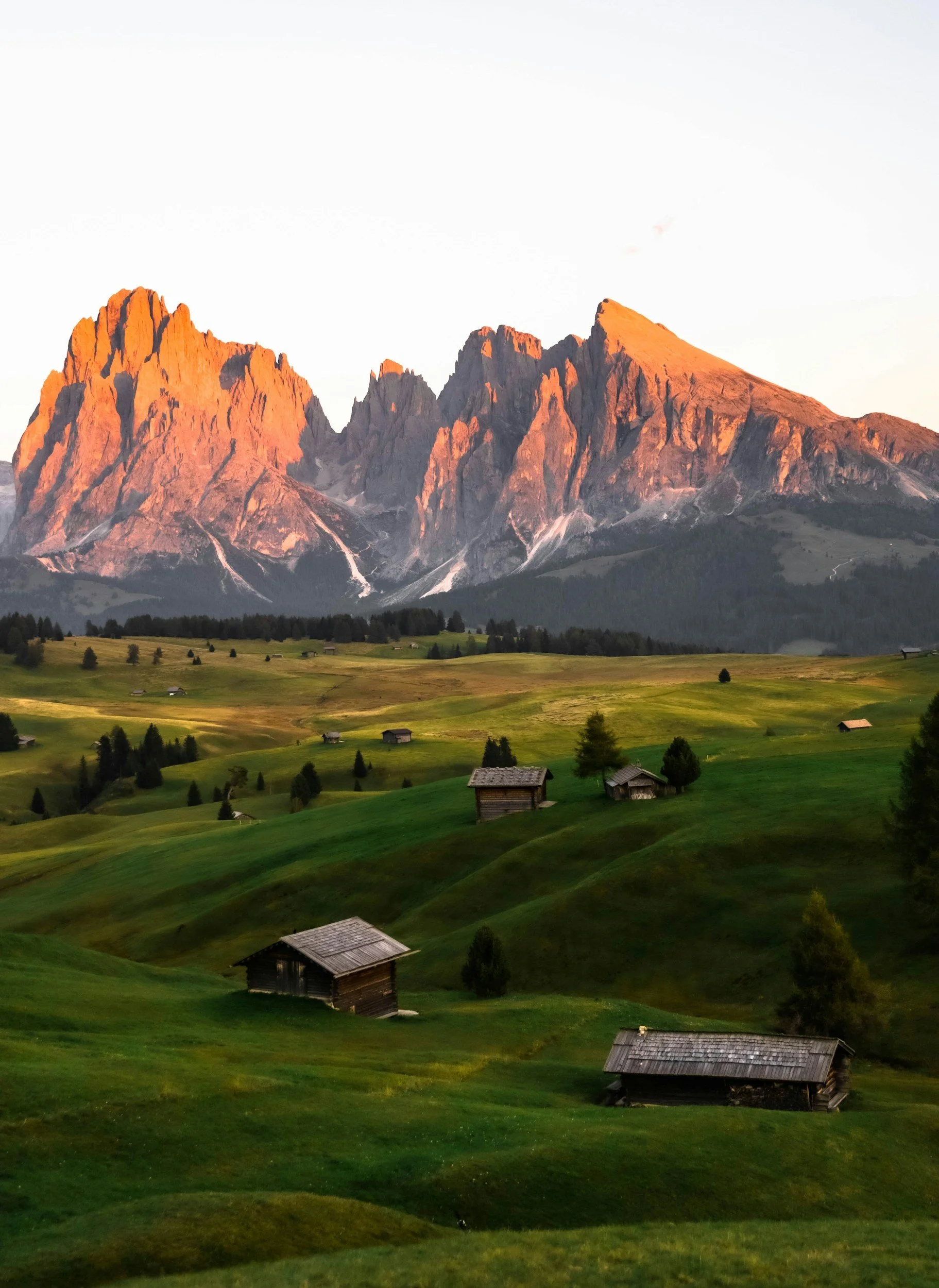 A landscape of green rolling hills and small wooden houses in front of a mountain range with rocky peaks bathed in sunset light.