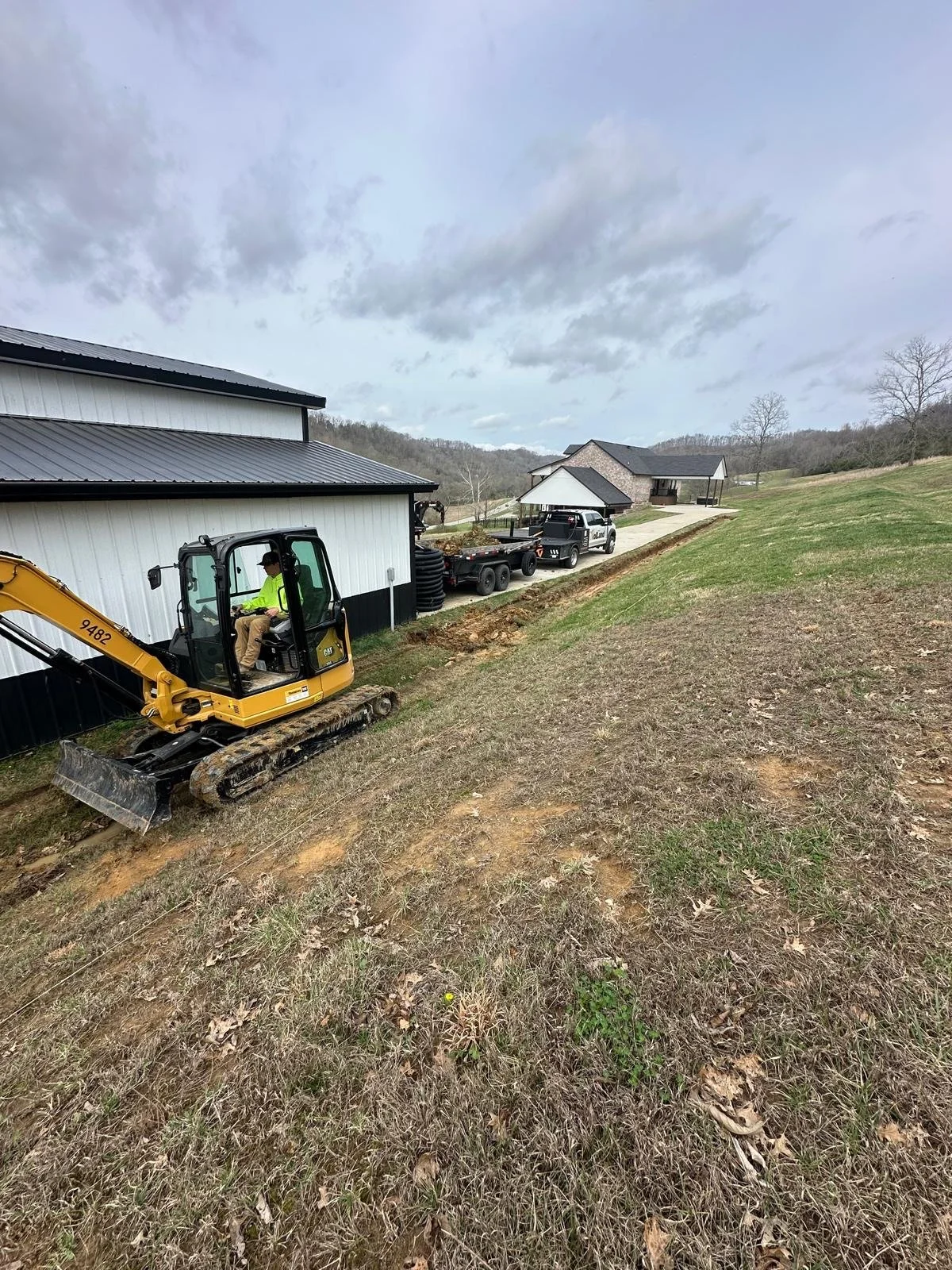 A construction site with a yellow excavator working on a grassy slope, and a white and black building to the left. There are two pickup trucks parked nearby, and residential houses in the background under a cloudy sky.