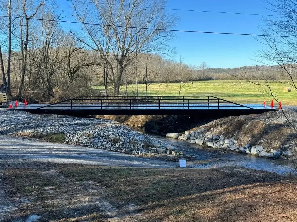 A small bridge over a creek with construction cones and equipment nearby, in a rural area with trees and hay bales in the background.