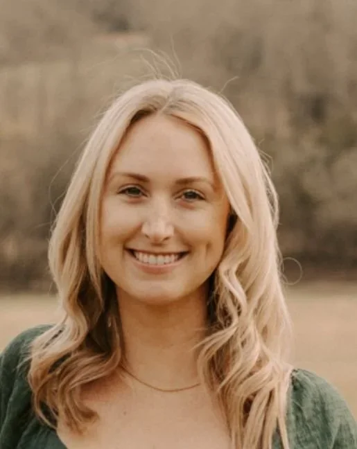 A young woman with long blonde hair smiling outdoors in a natural setting.