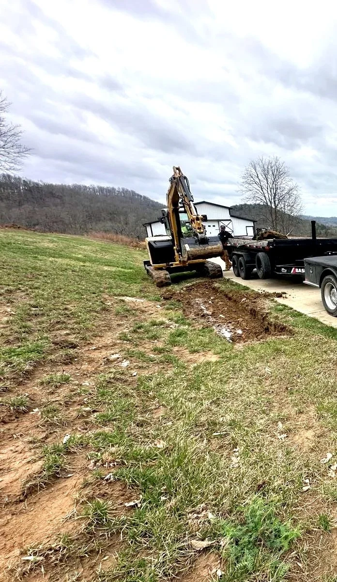 A small excavator digging a trench near a grassy hill, with a flatbed trailer and a white building in the background.