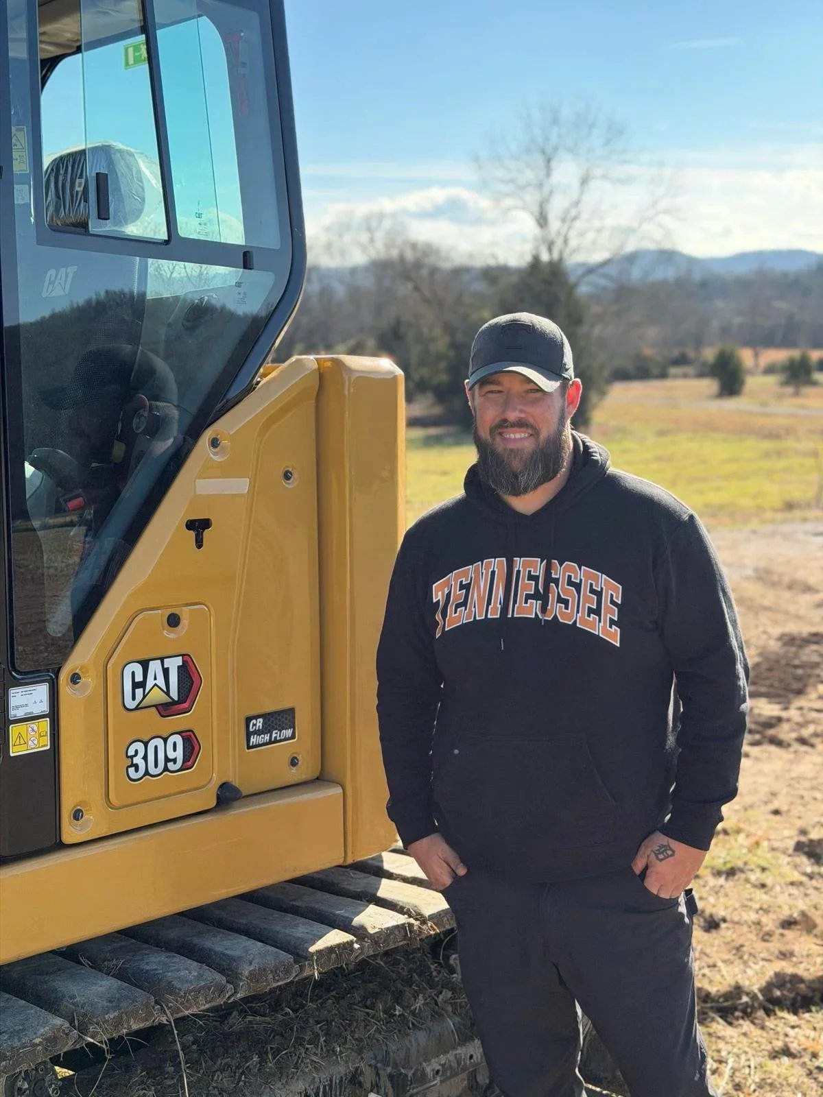 A man with a beard wearing a Tennessee hoodie and cap standing outdoors next to a yellow Caterpillar 309 excavator, with trees and mountains in the background on a sunny day.