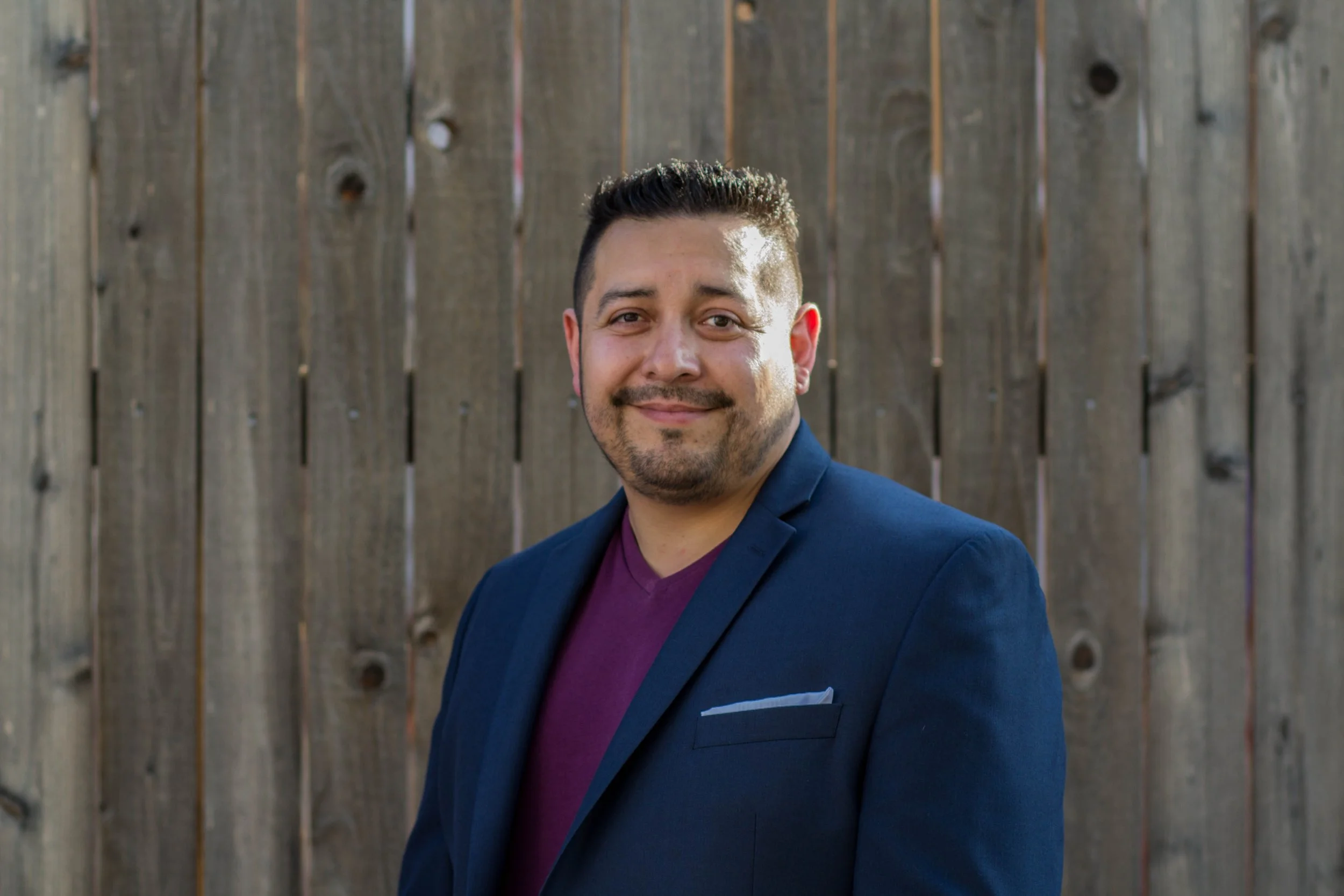 A man with short dark hair and a beard, wearing a navy blazer and a maroon shirt, standing in front of a wooden fence, smiling at the camera.