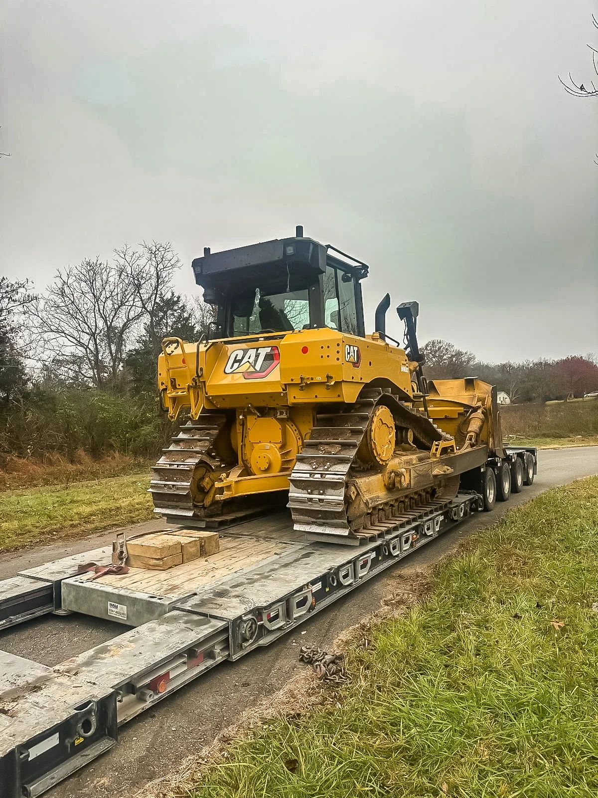 A yellow Caterpillar bulldozer is being transported on a flatbed trailer along a rural road on an overcast day.