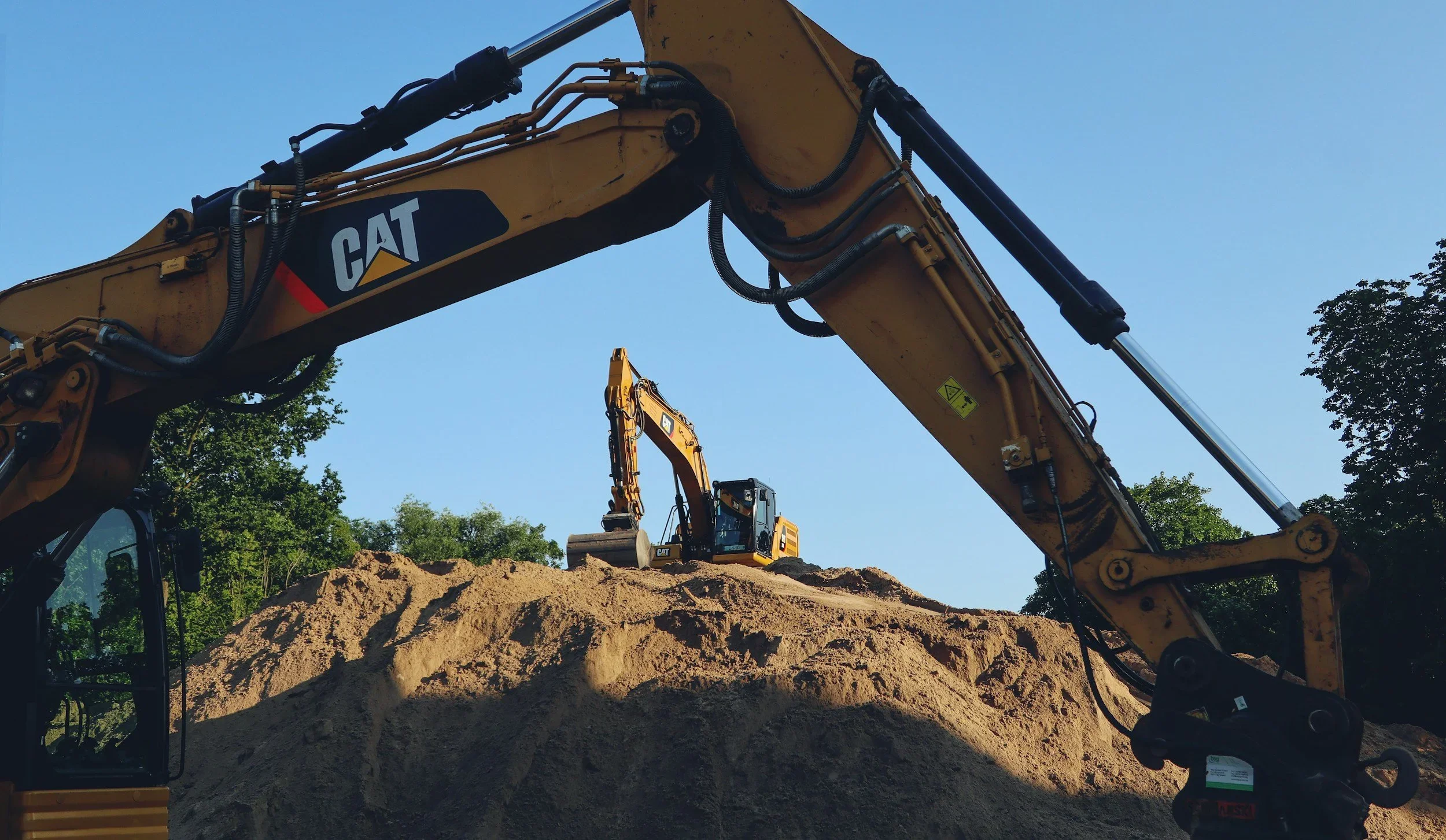 Construction site with a large yellow Caterpillar excavator digging into a mound of dirt against a blue sky with some trees in the background.