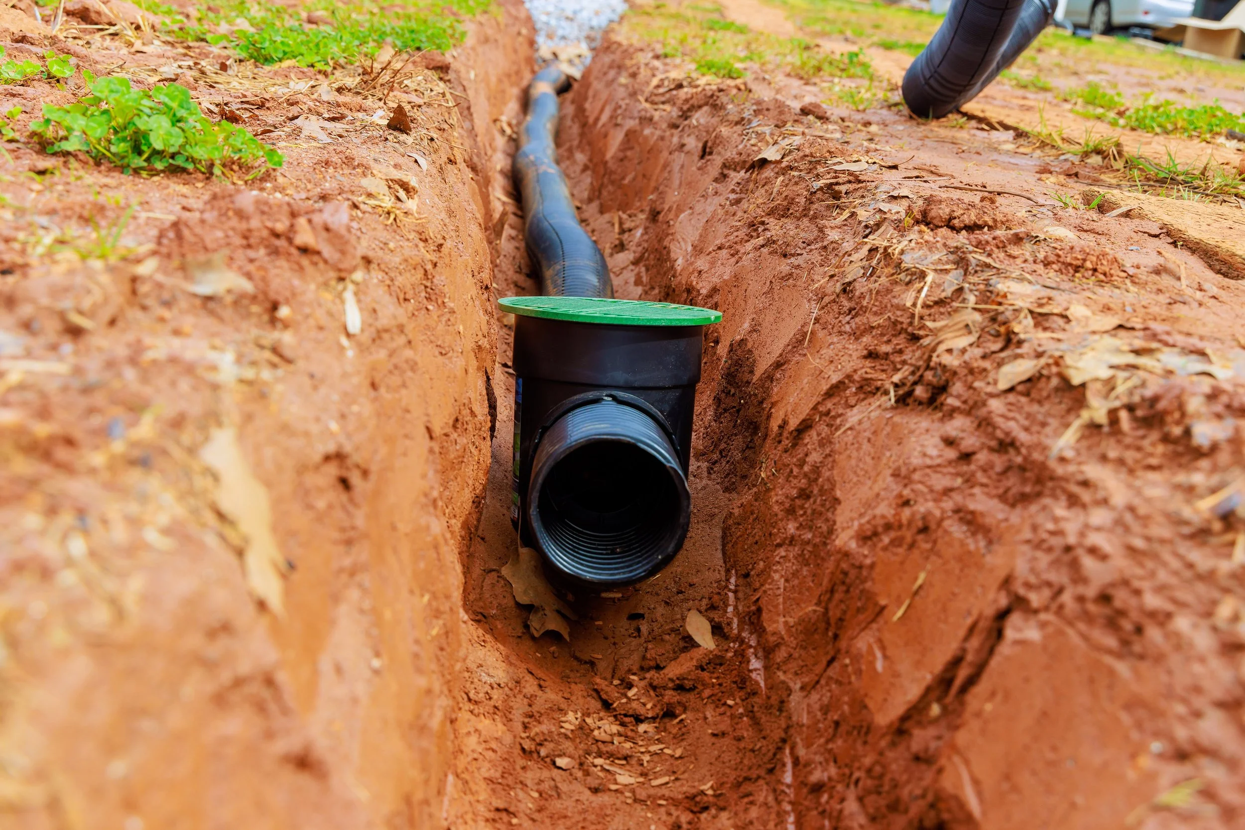 A black underground drainage pipe in a trench in red dirt, with a green cover on top, connected to a larger pipe extending into the trench, with some small green plants on the side.
