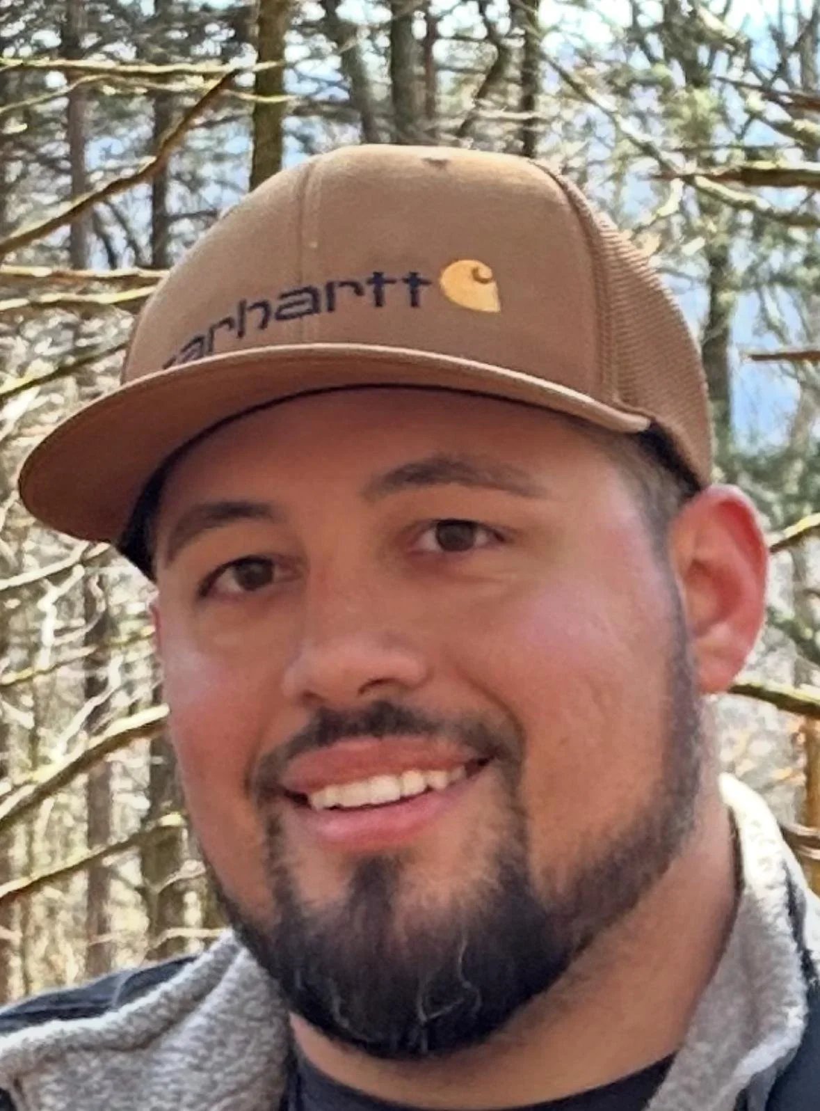 Close-up of a smiling man wearing a tan Carhartt baseball cap outdoors with trees in the background.