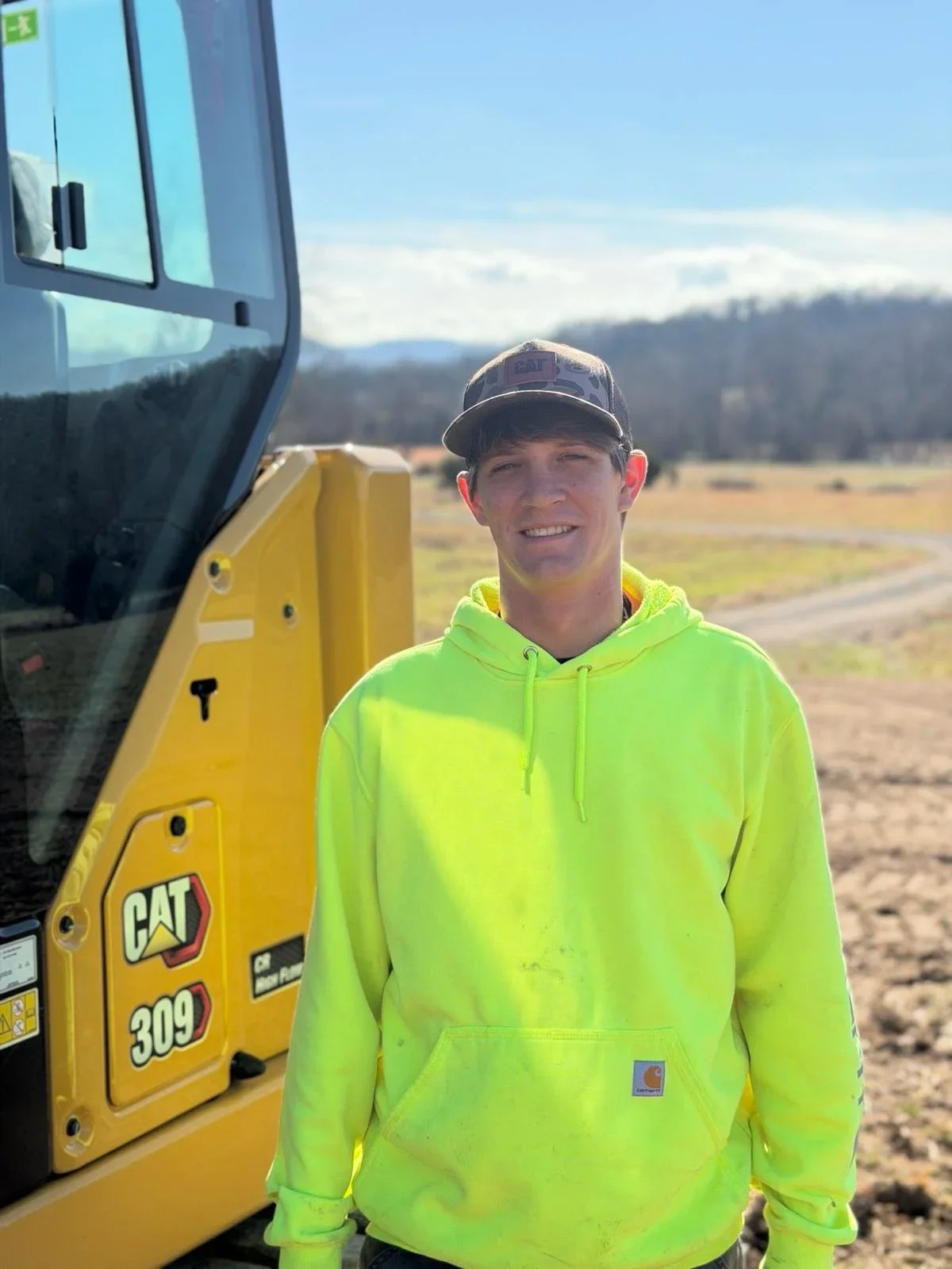 A young man wearing a bright yellow hoodie standing outdoors next to a yellow Caterpillar construction vehicle on a dirt field with mountains and a cloudy sky in the background.