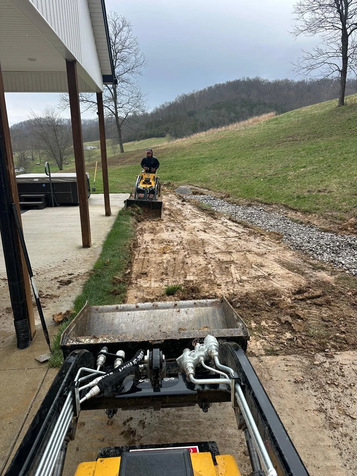 View from a construction vehicle looking at a worker using a bulldozer to level a dirt path outside a house, with trees and hills in the background.