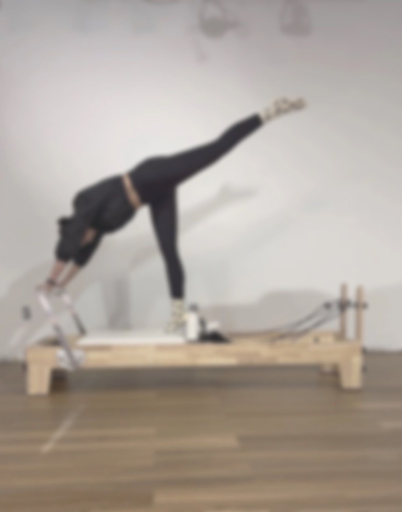 A person doing an arebesque on a wooden reformer machine in a room with a white wall and wooden floor.