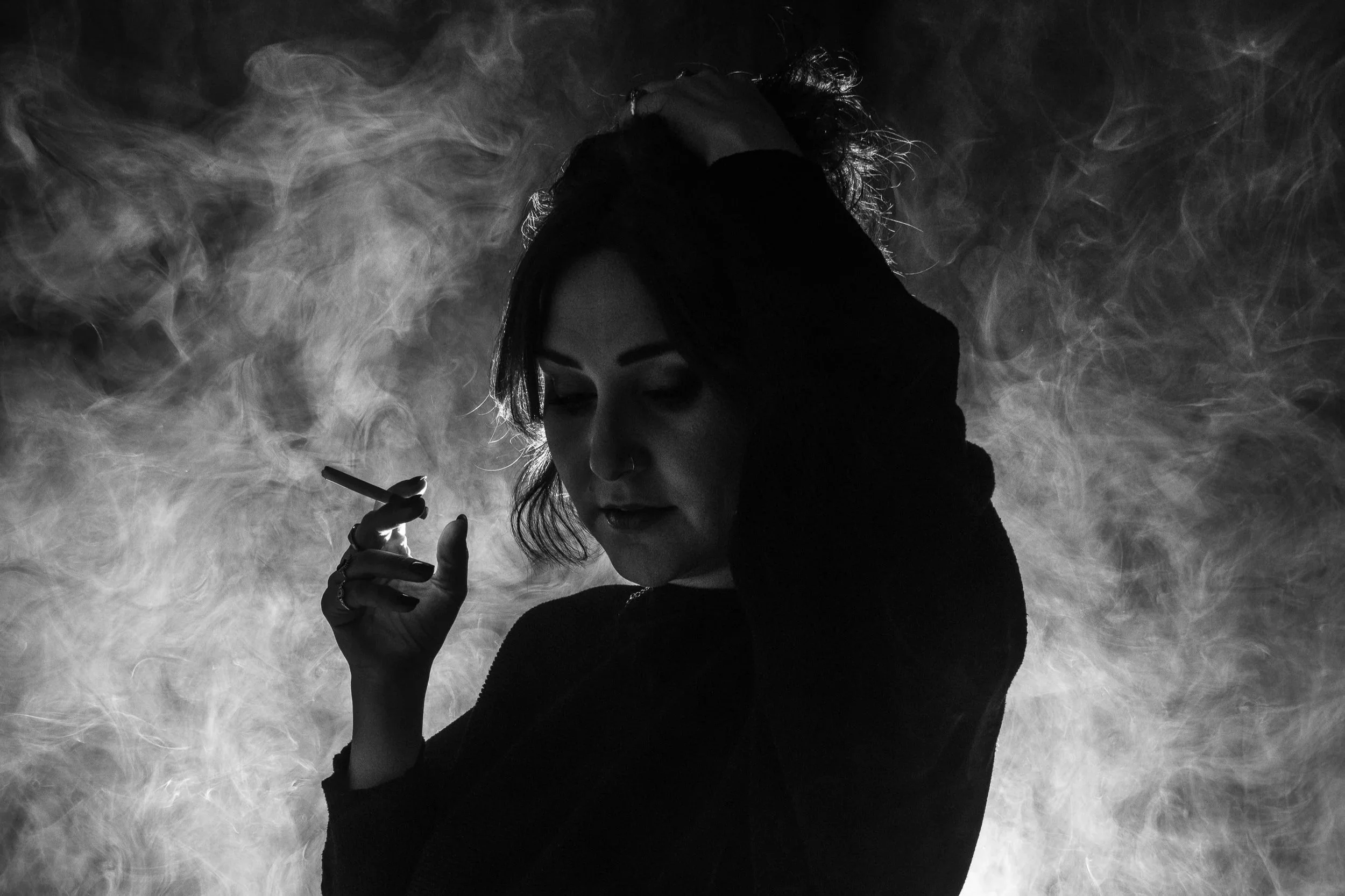 A black and white photo of a woman with curly hair holding a cigarette, with smoke swirling around her head against a dark background.