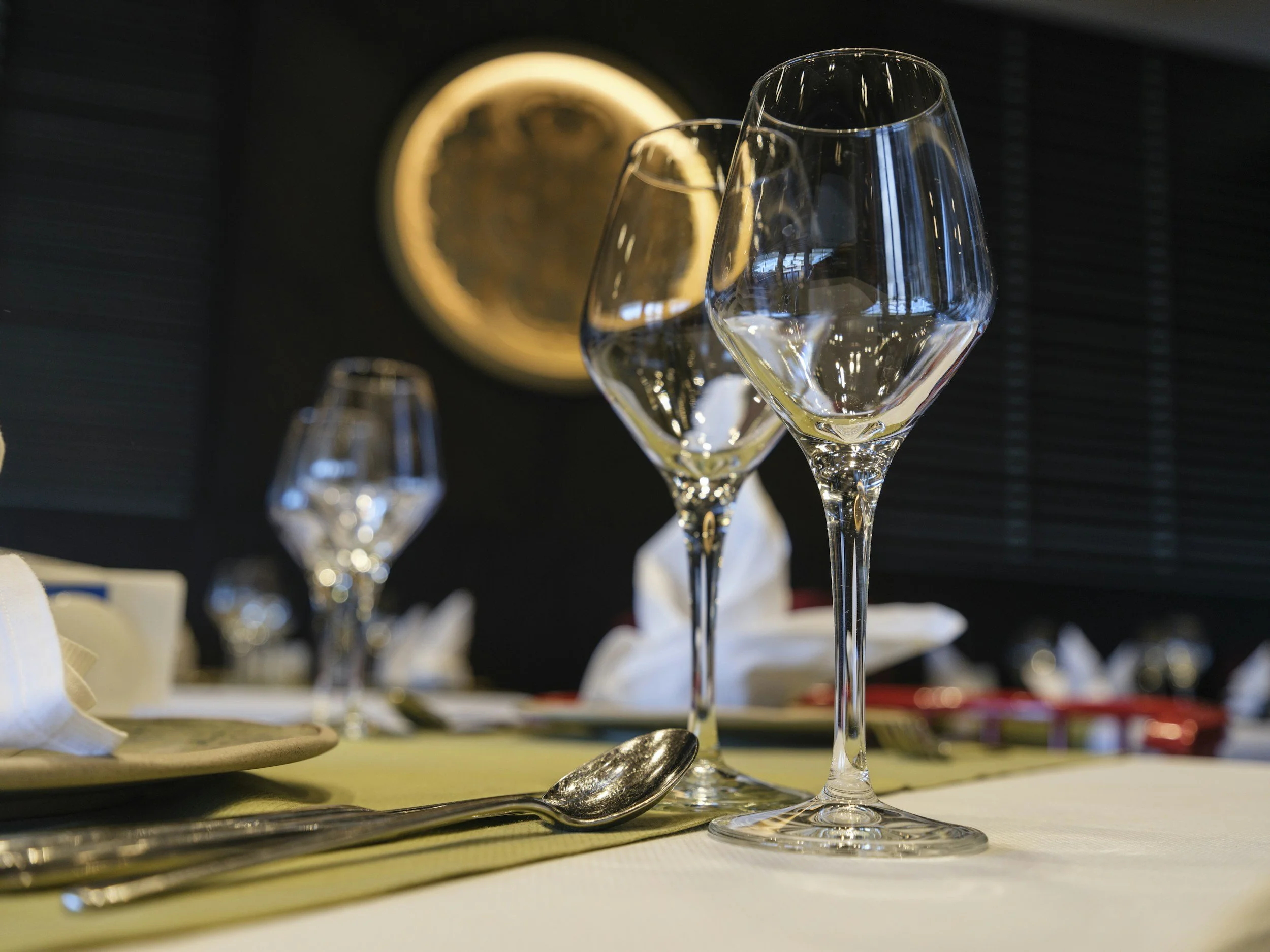Empty wine glasses on a table setting with a spoon and napkin, in a dimly lit room with dark walls and a circular art piece in the background.