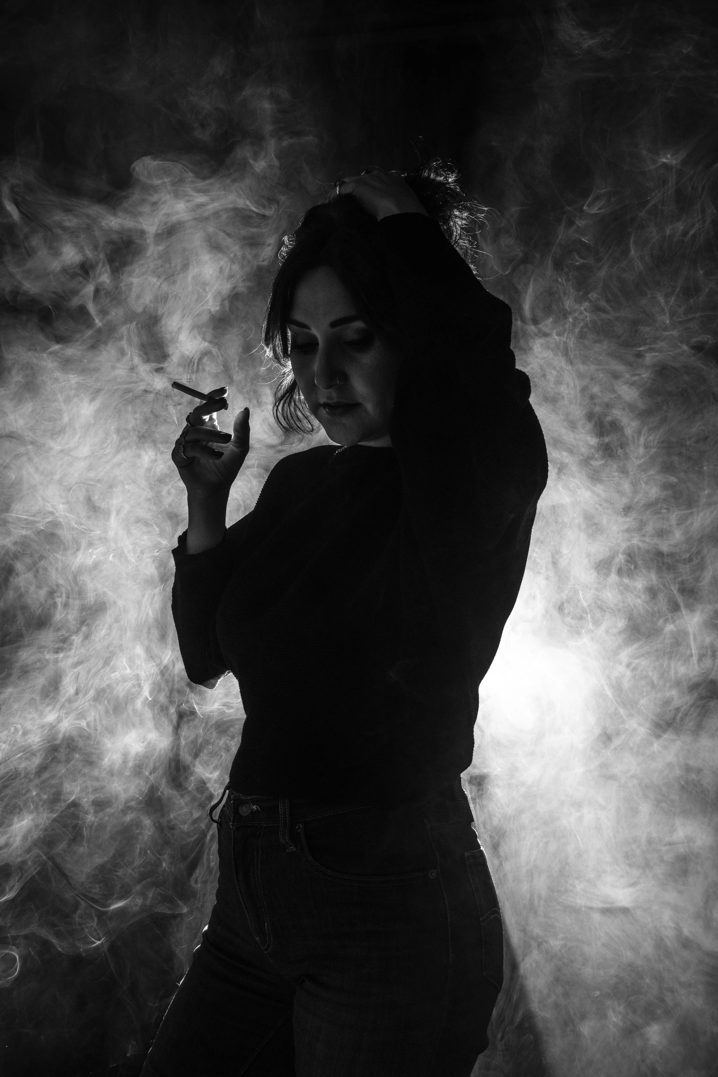 A black and white photo of a woman with dark hair, standing among swirling smoke or fog, holding a cigarette near her face, with her head tilted down and one hand on her head.