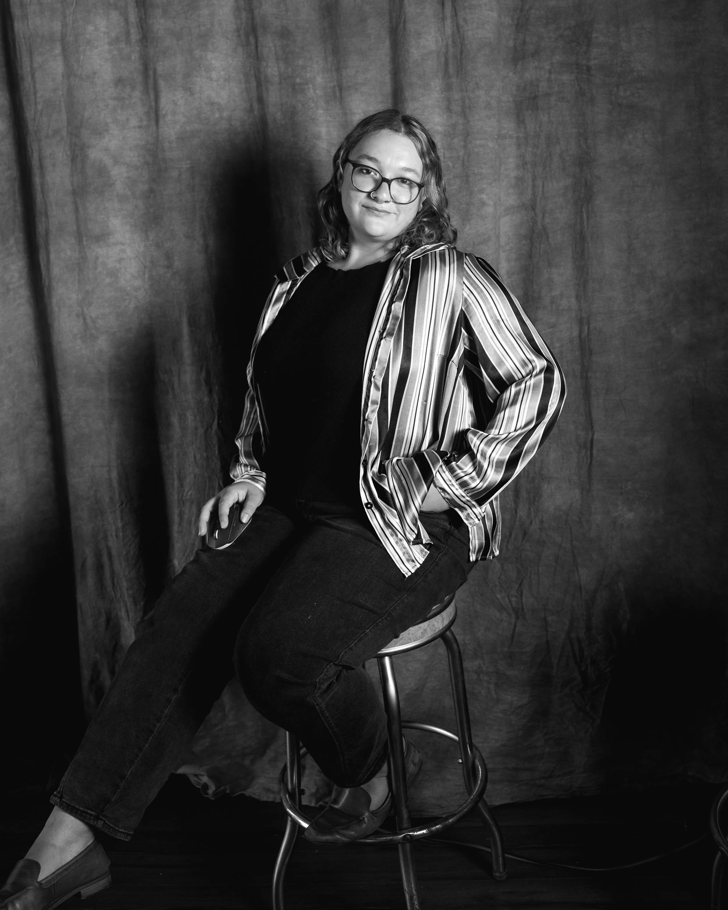 A person with glasses and a nose ring sitting on a stool in front of a dark curtain, wearing a striped shirt and dark jeans holding a phone.