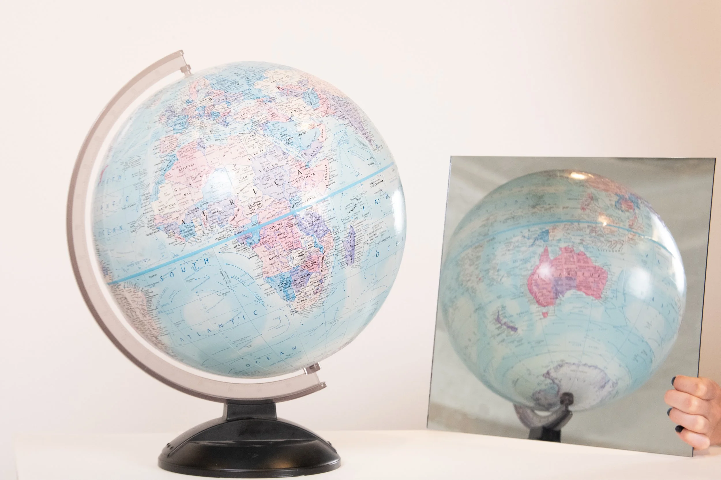 A globe and a mirror reflecting the globe, with a person's hand holding a small globe. The globe displays Africa, Europe, and parts of Asia.