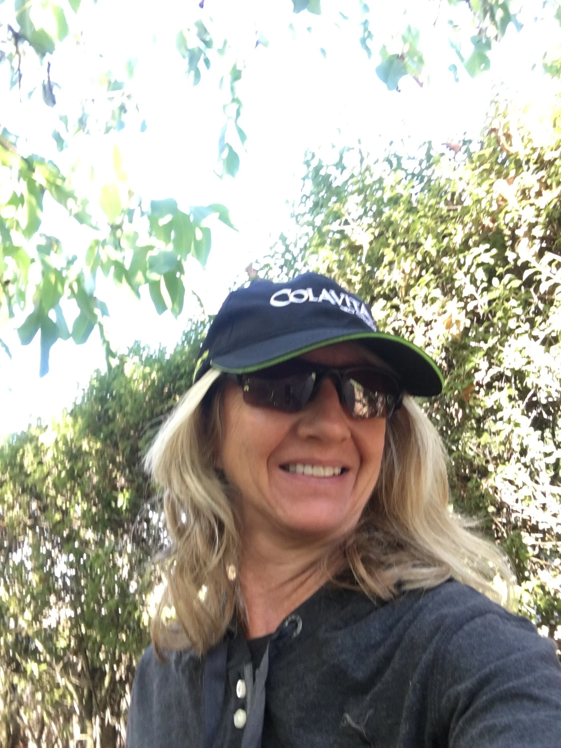 A woman with blonde hair and sunglasses, smiling, wearing a navy cap with 'COLAVITA' on it and a black jacket, stands outdoors among trees and greenery.