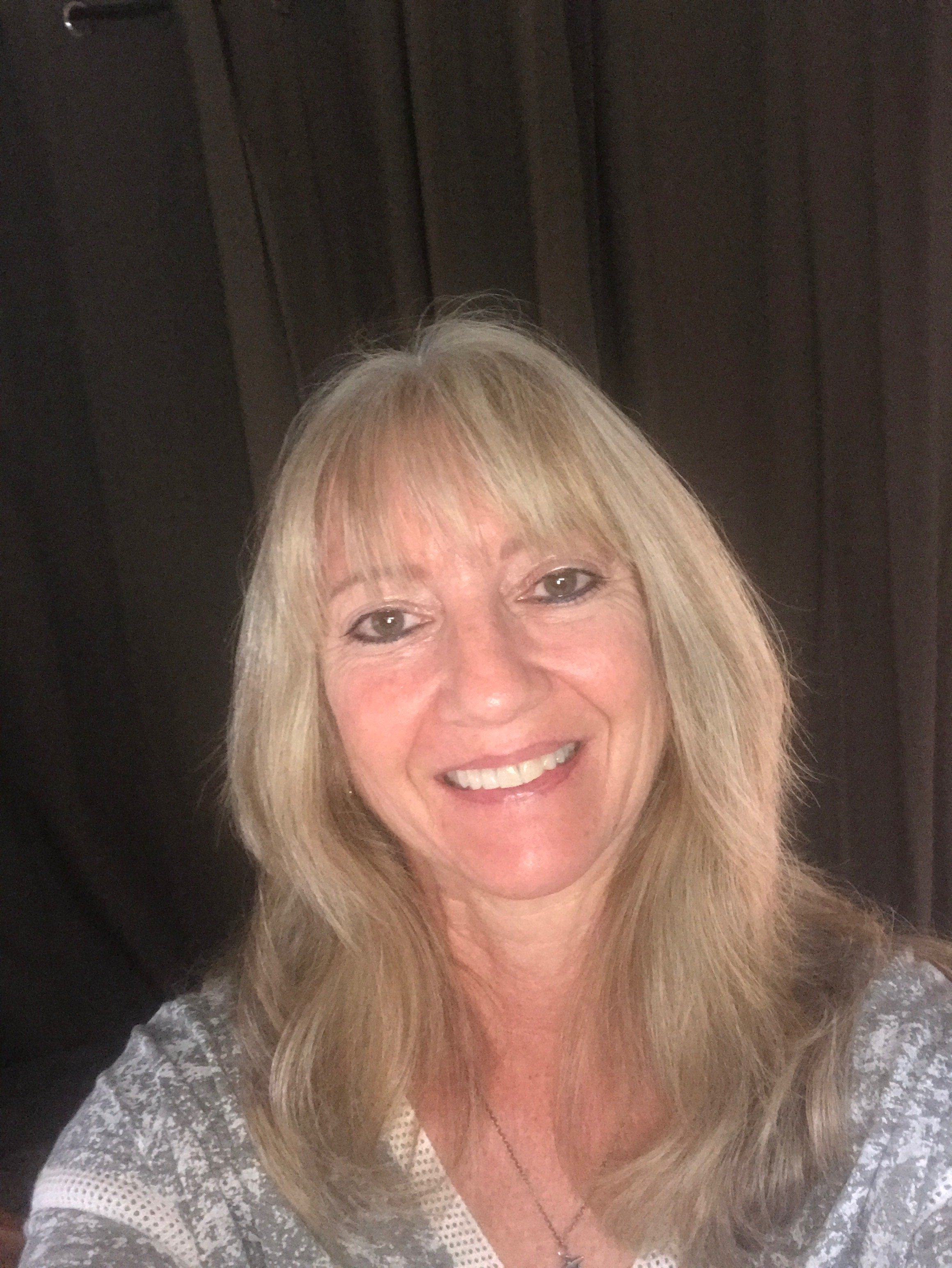 A smiling woman with long blonde hair, wearing a light-colored lace top and a necklace, standing against a dark wooden background.