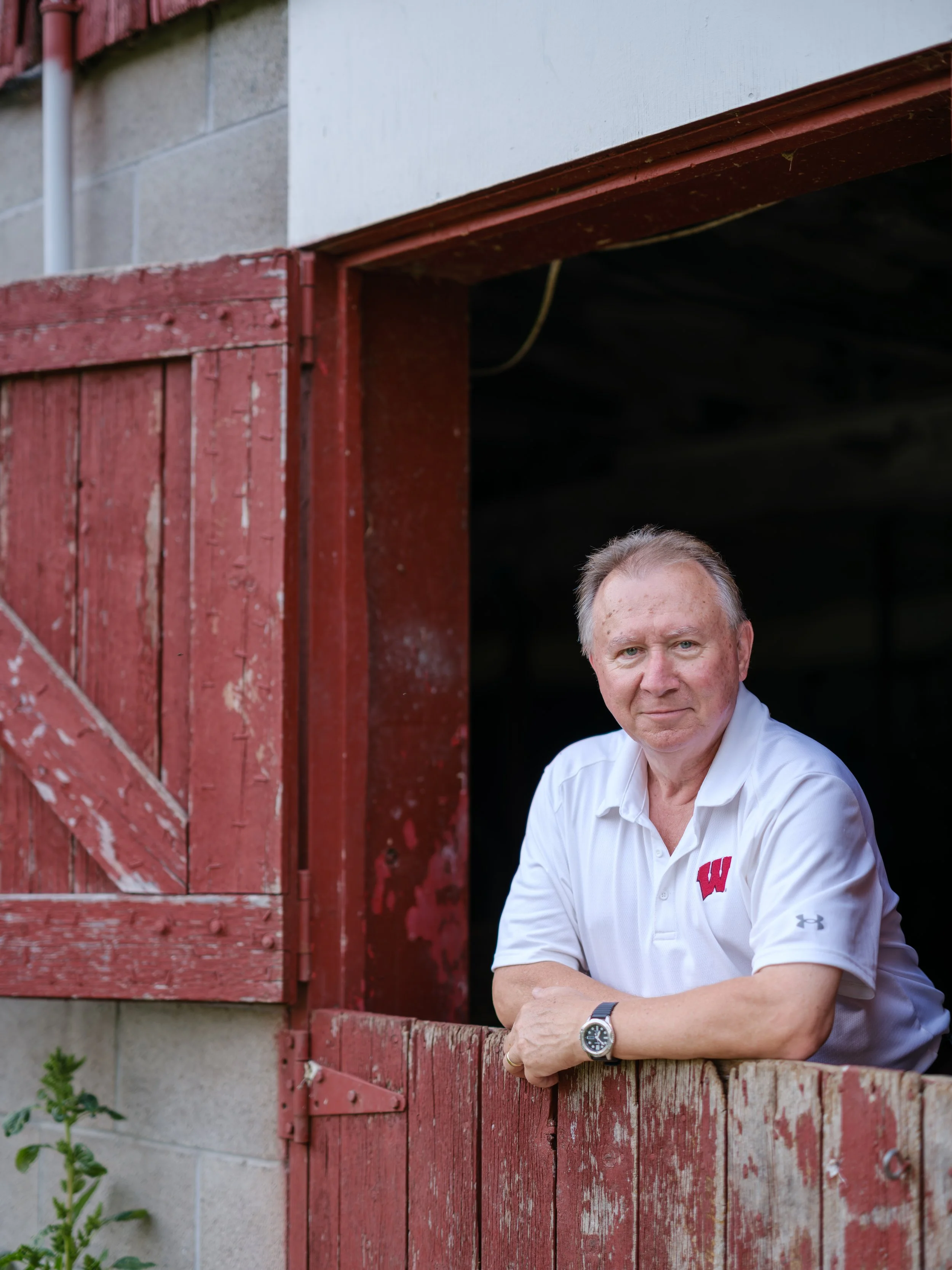 State Representative Steve Doyle leans on a weathered red barn window frame, wearing a white shirt with a red 'W' logo and an Under Armour logo on the sleeve, and a wristwatch, looking at the camera.
