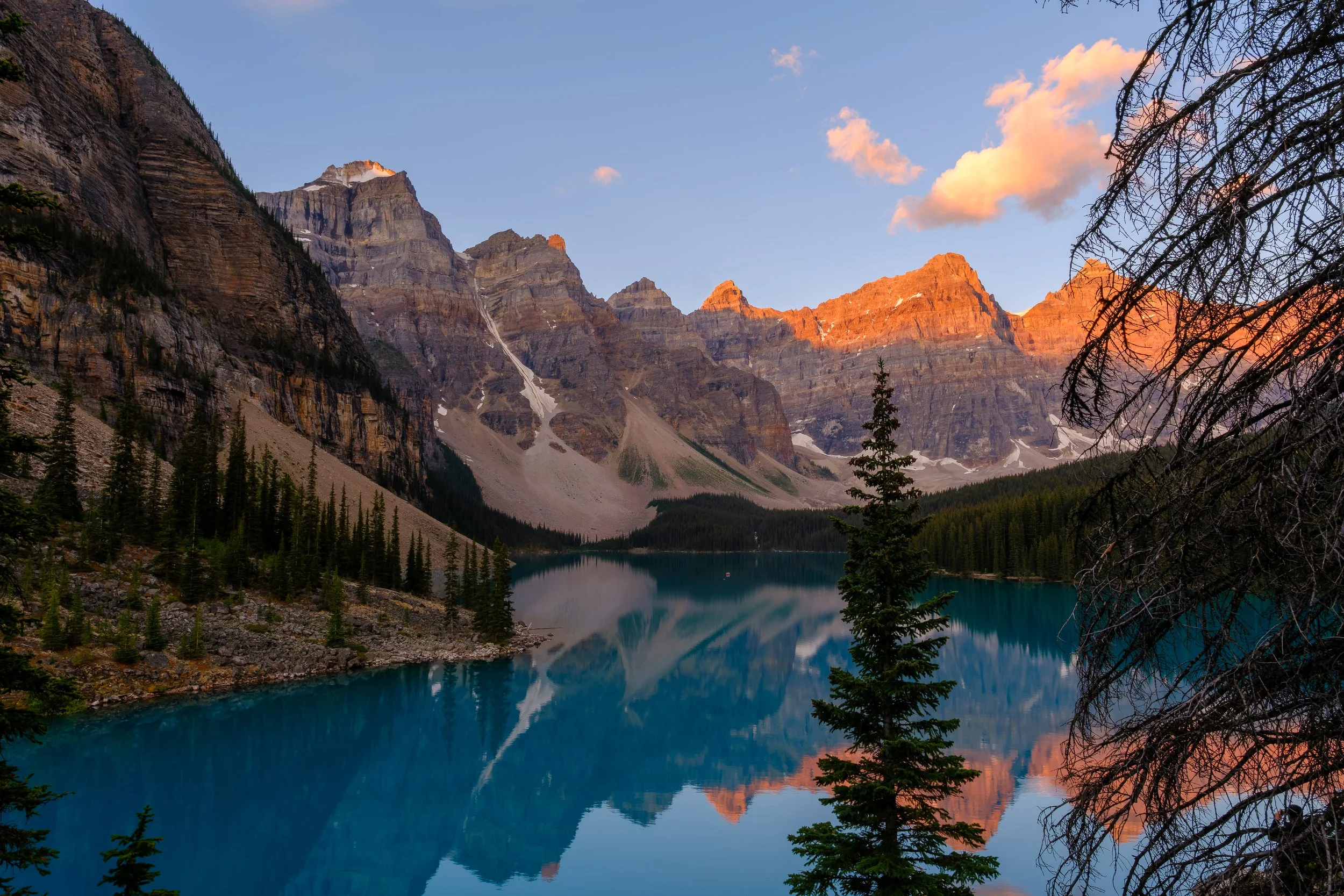 Scenic mountain landscape with a calm, reflective lake, surrounded by evergreen trees, with rugged snow-capped peaks illuminated by the rising sun, and a partly cloudy sky from Moraine Lake in Banff National Park.