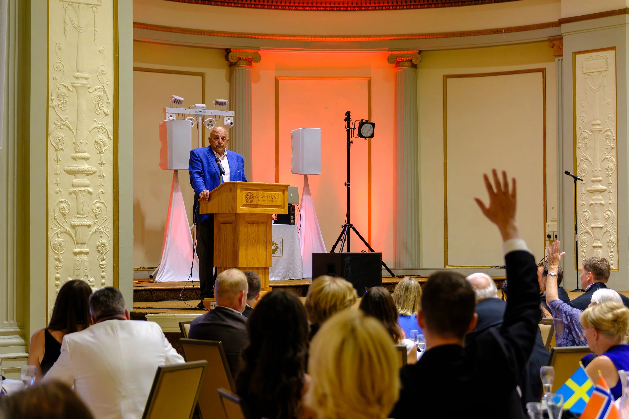 Barry Alvarez speaks at a podium during an event, with audience members seated in front, some raising their hands, inside an ornate hall with decorative wall moldings and lighting.