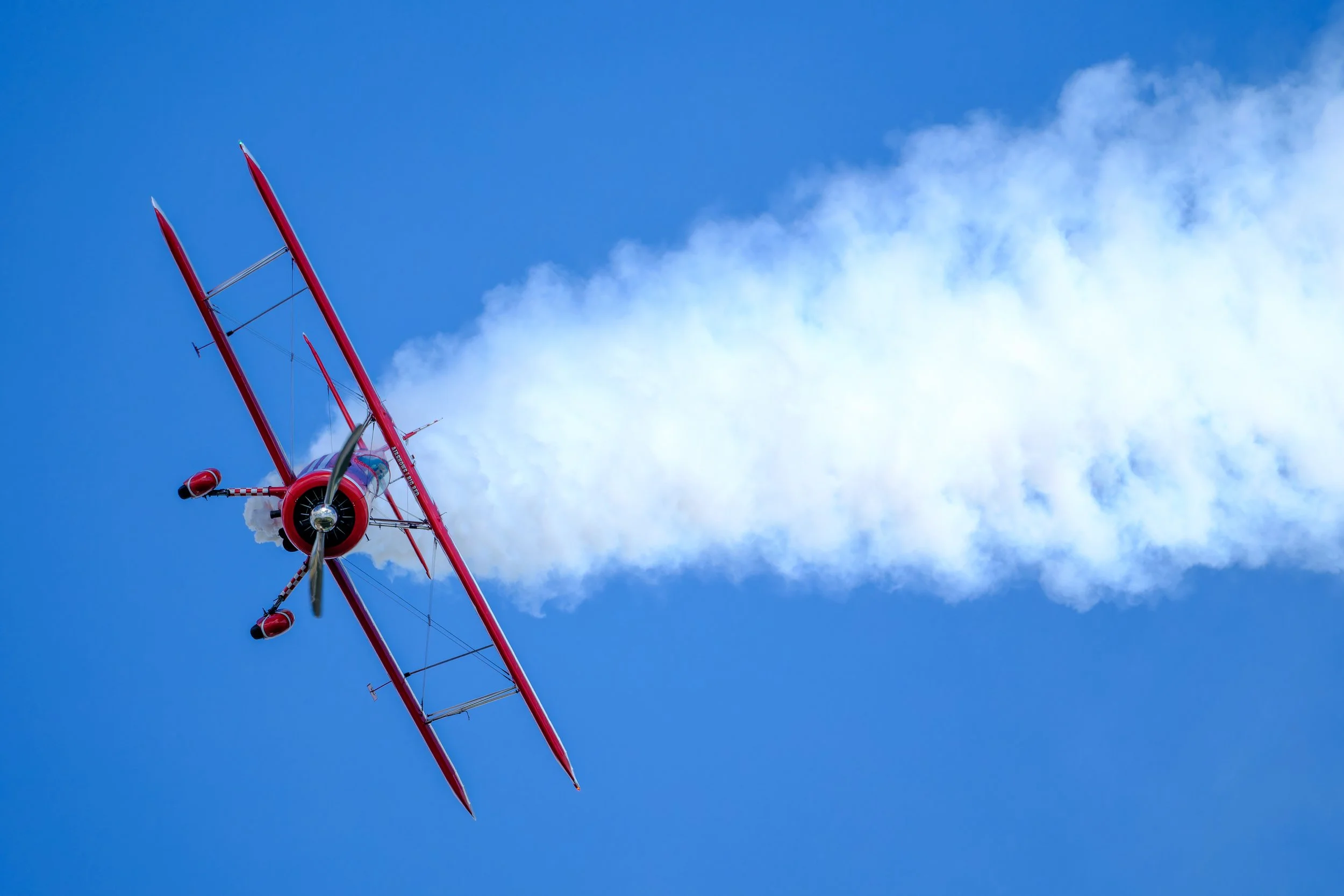 Red aerobatic biplane performing an inverted maneuver, leaving a trail of white smoke against a blue sky. From EAA AirVenture in Oshkosh, Wisconsin.