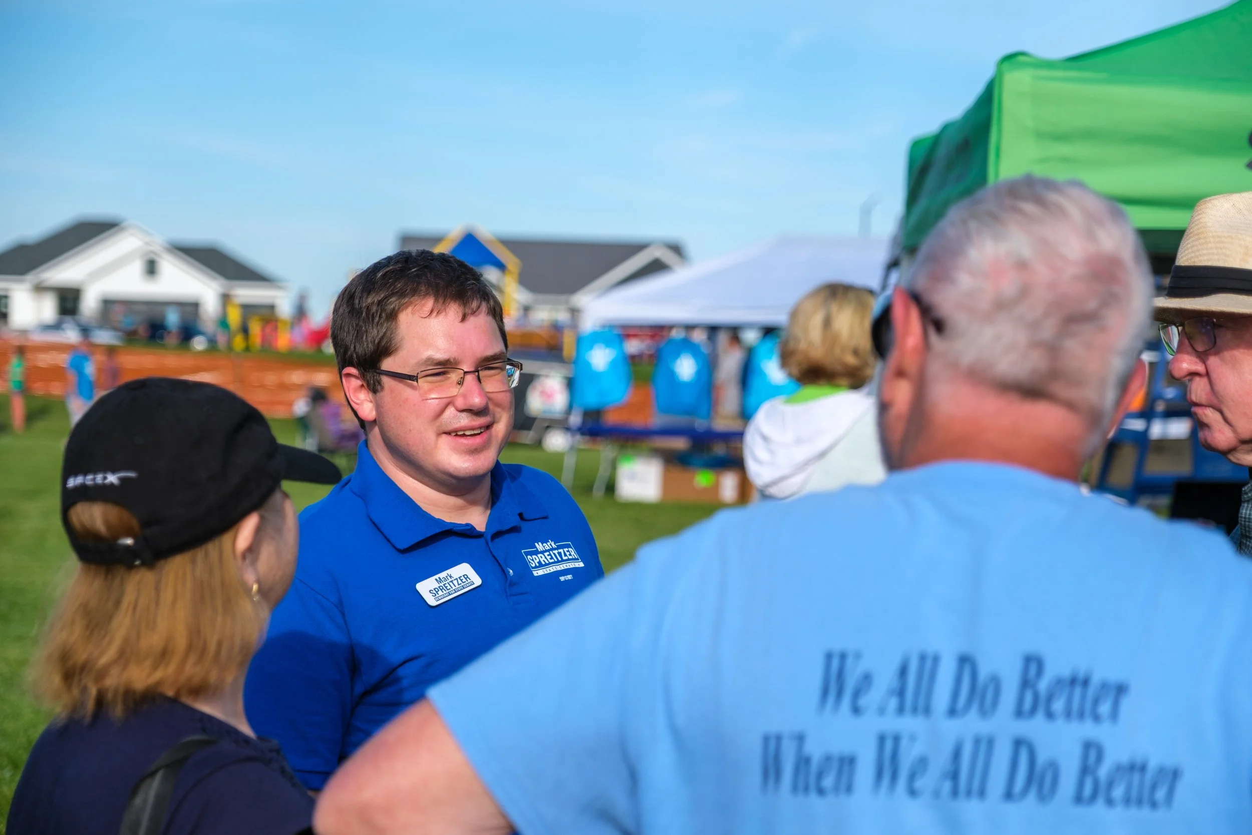 Group of people talking outdoors at a fair or festival, with tents, booths, and houses in the background.