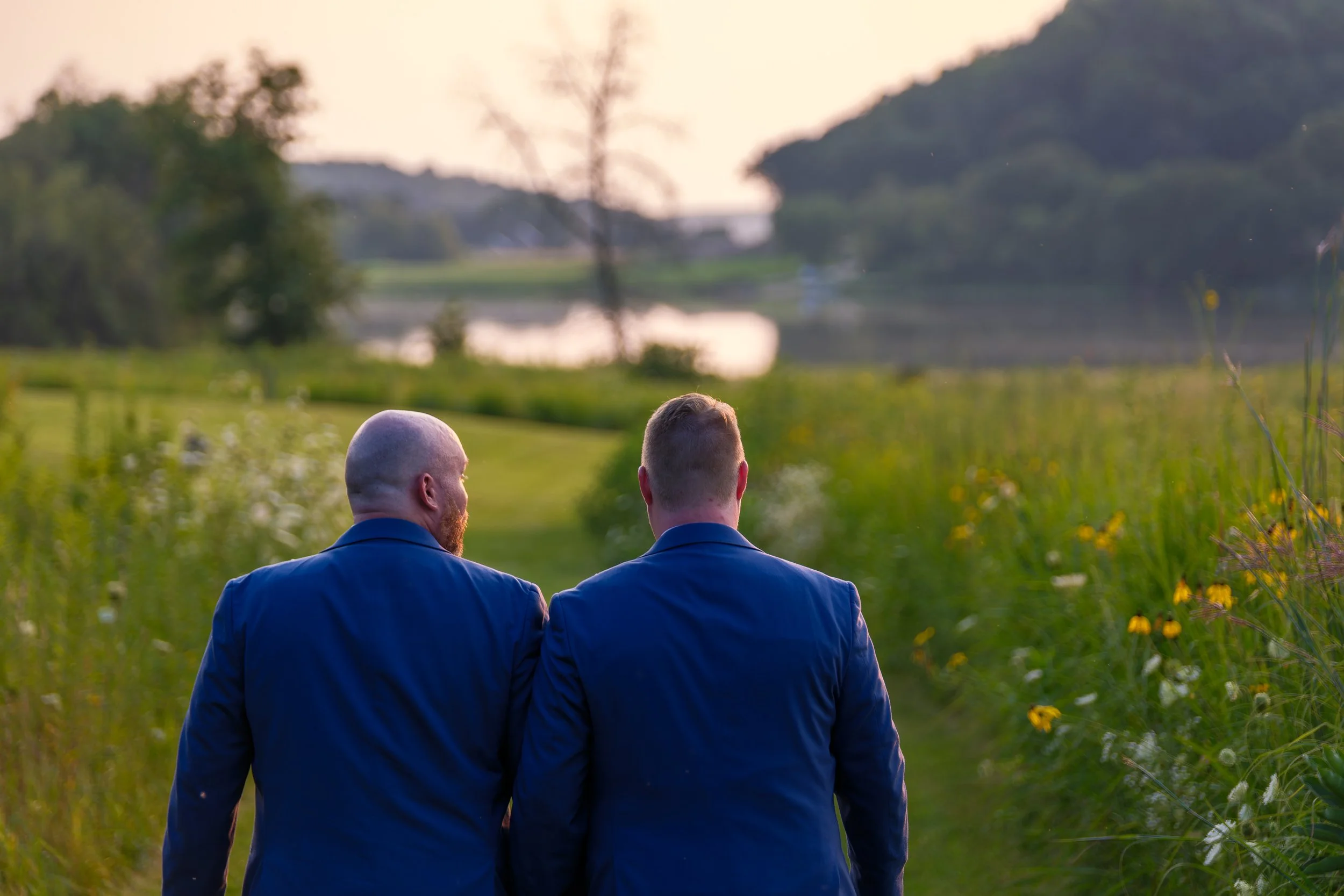 Two men in blue suits walking through a lush green field toward a lake at sunset, with trees and hills in the background.