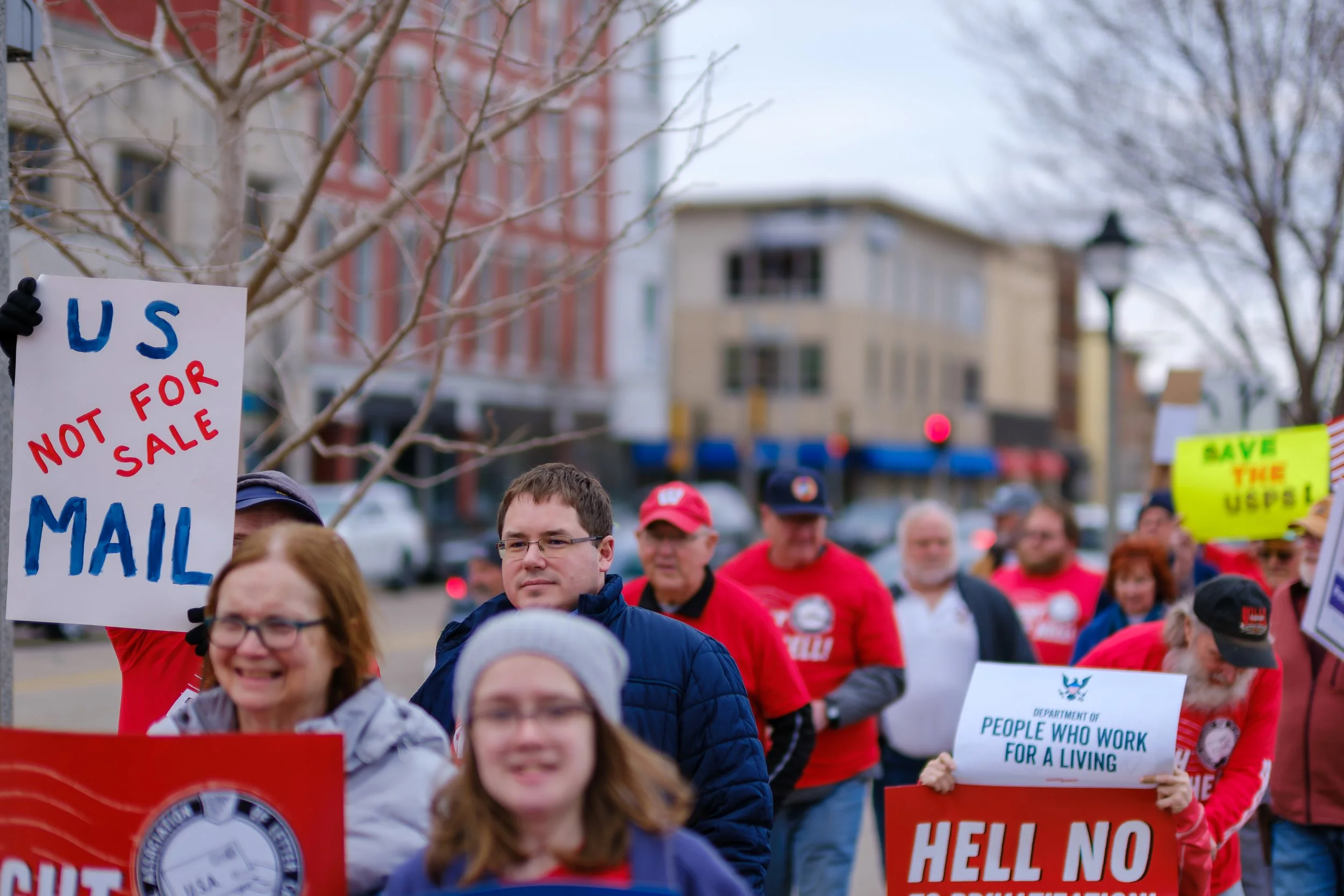 Protest march with people holding signs including one that says 'US MAIL NOT FOR SALE' and another that says 'SAVE THE USPS'. The protesters are walking down a city street with buildings and leafless trees in the background, wearing casual clothing a