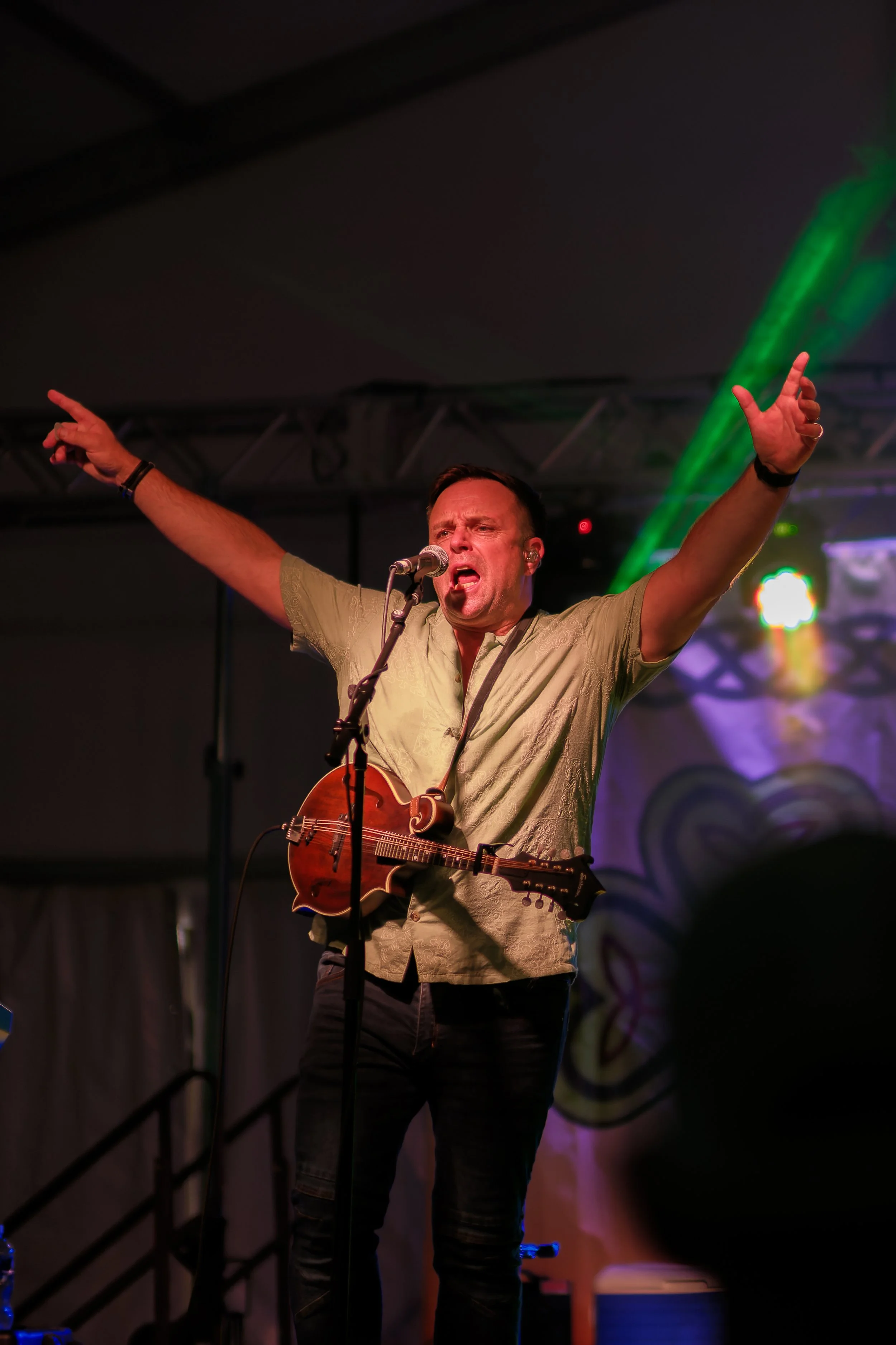 Male musician performing on stage with a microphone and acoustic guitar, arms raised, in a concert setting with colorful lights.