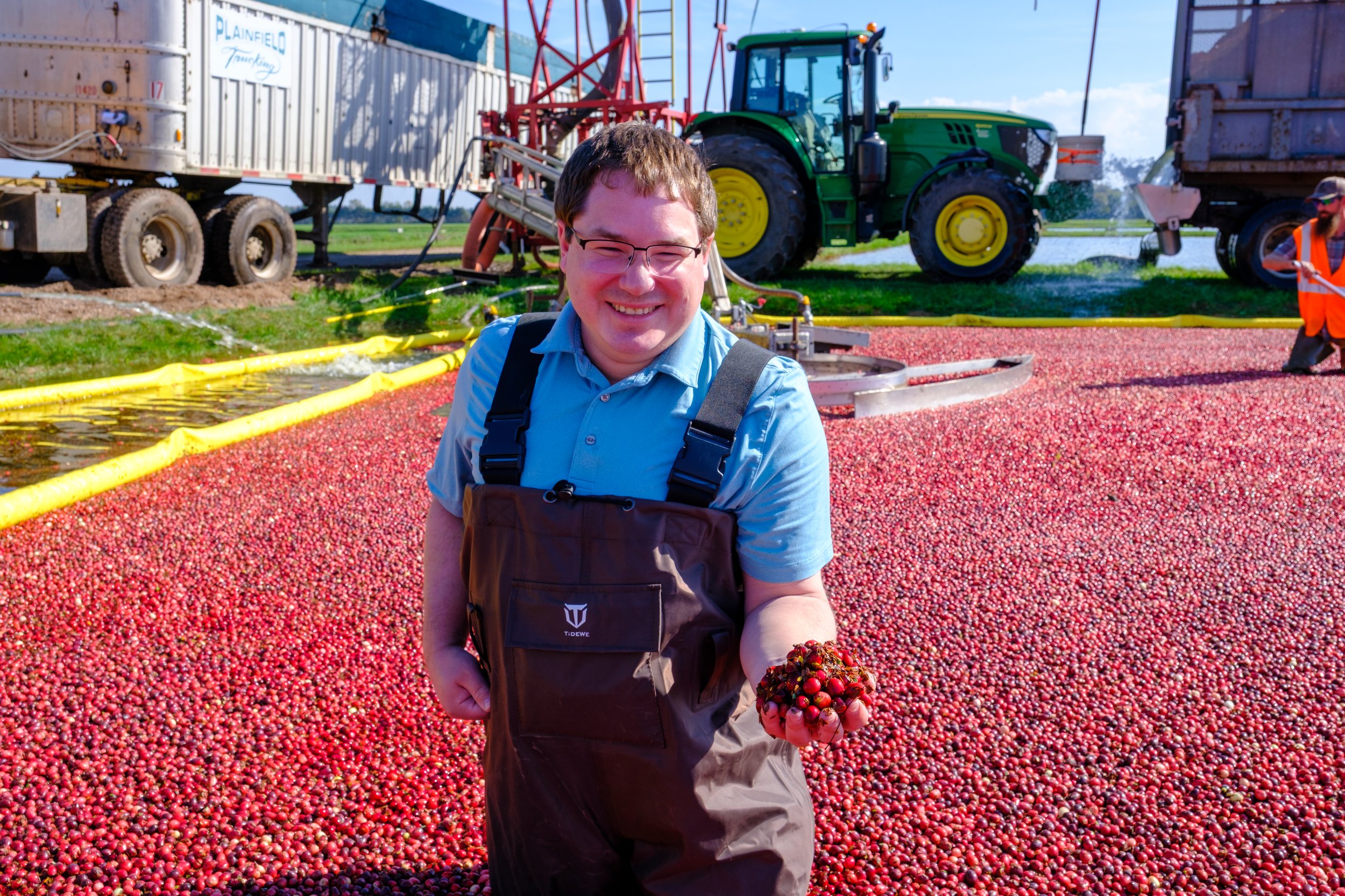 Wisconsin State Senator Mark Spreitzer holds a handful of cranberries in a flooded cranberry marsh. Background shows a large number of cranberries, harvesting equipment, and a tractor.