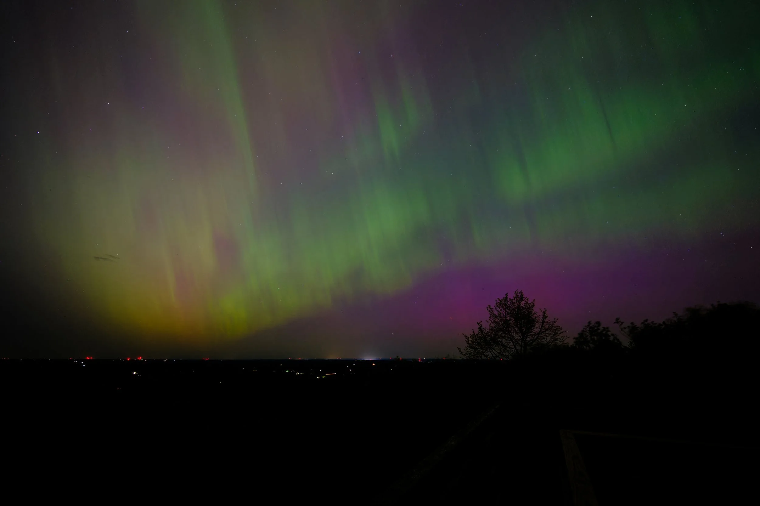 Northern lights glowing green, purple, and yellow in the night sky above trees and a distant landscape from Blue Mounds State Park in Wisconsin.