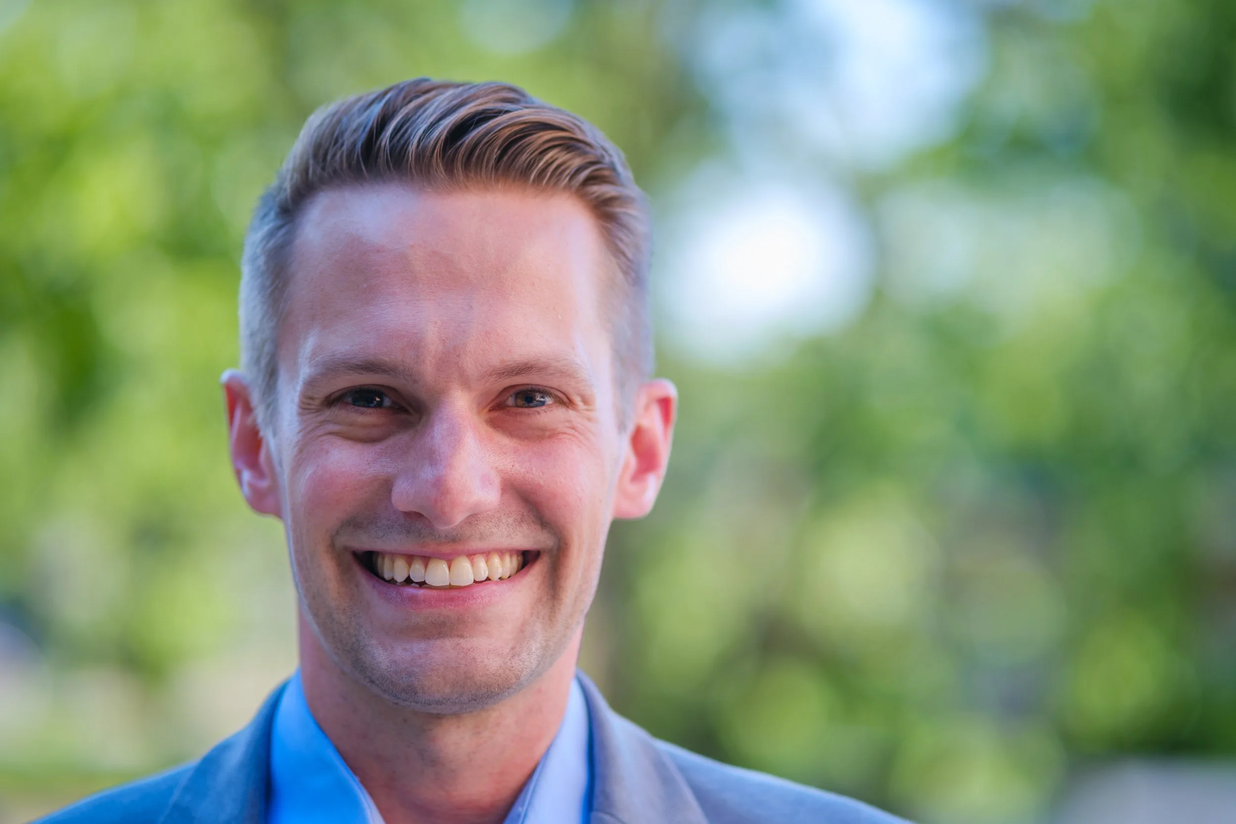 State Representative Alex Joers smiling outdoors with blurred green trees in the background.
