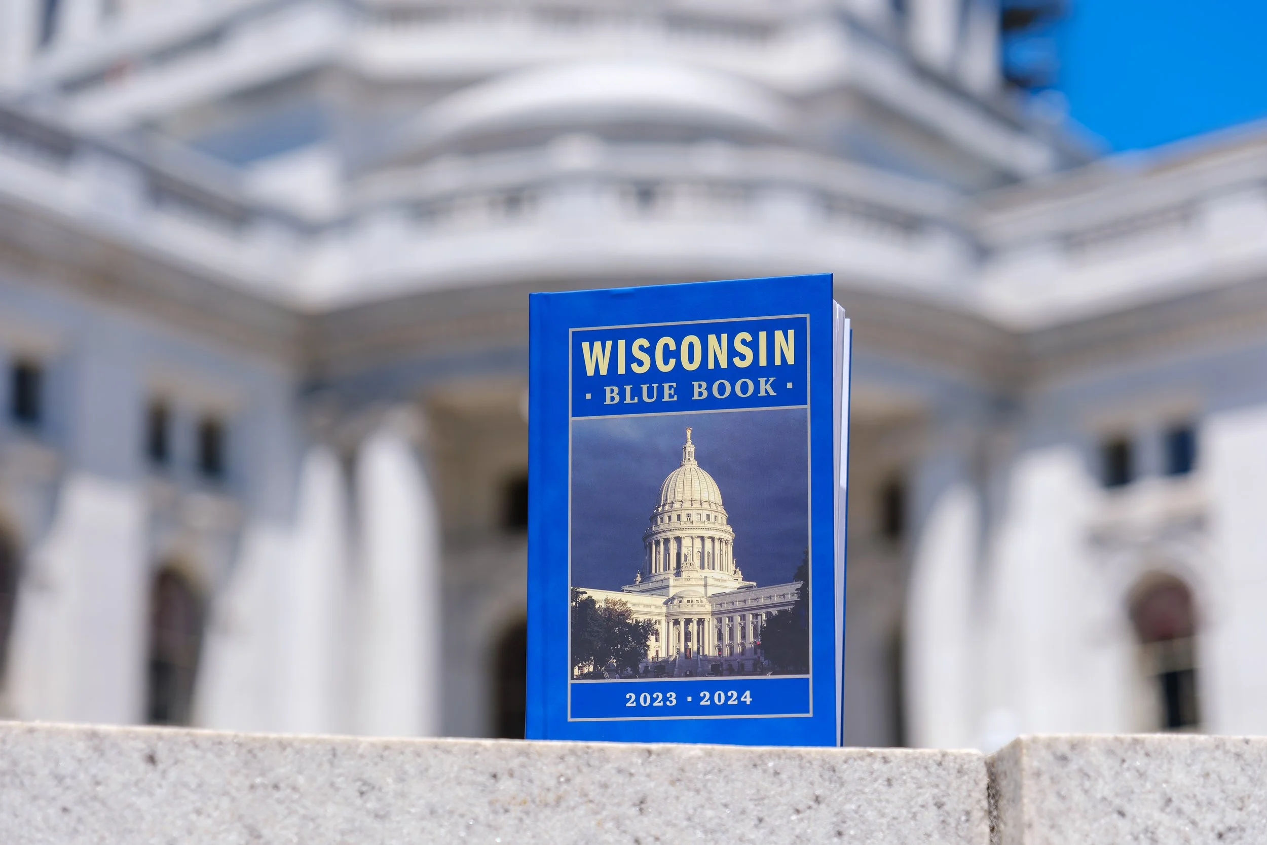 Blue Wisconsin Blue Book with an image of the Wisconsin State Capitol and text for the years 2023-2024, placed on a ledge in front of the Wisconsin State Capitol.
