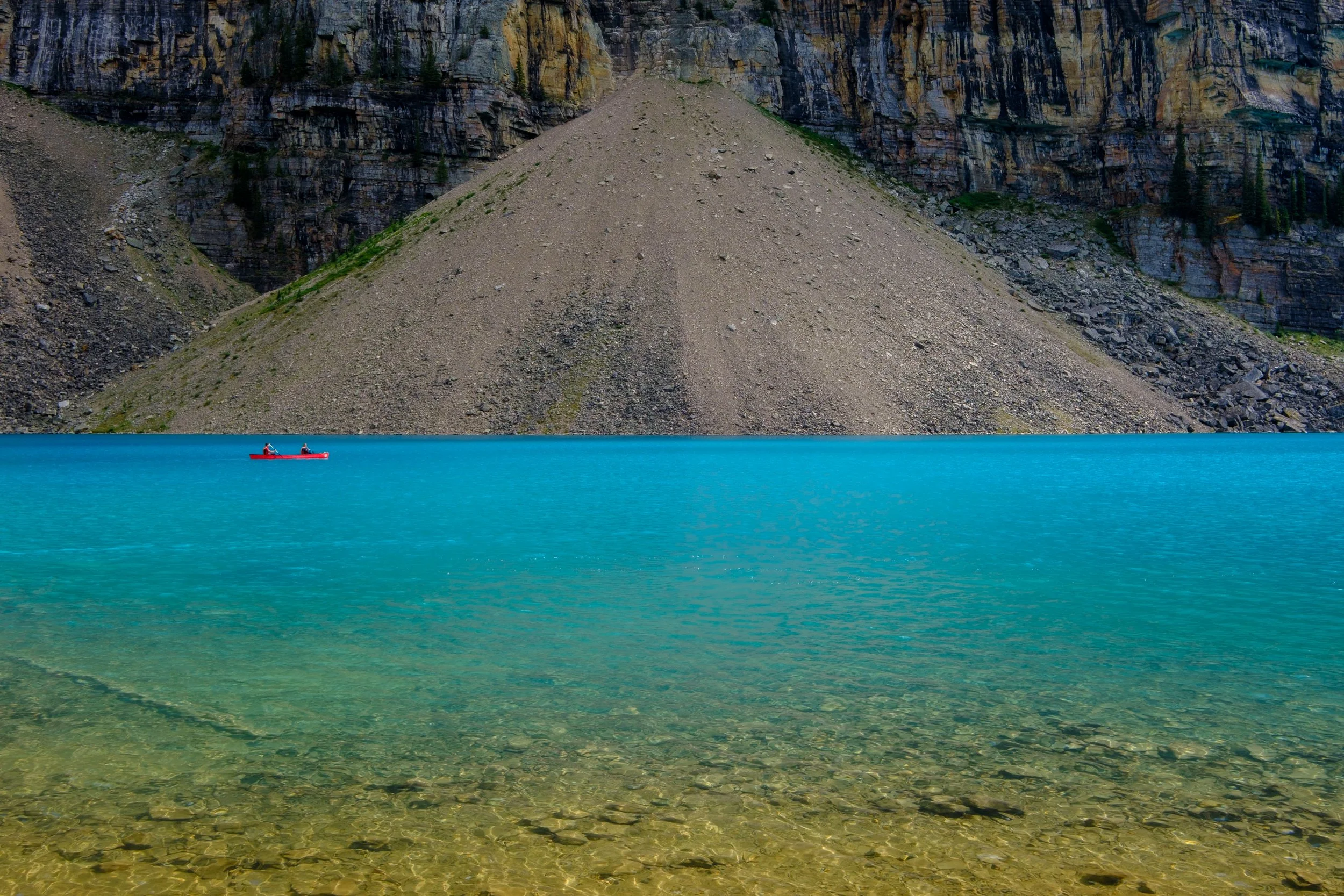 A bright blue Moraine Lake with a single red canoe and two people paddling, surrounded by rocky mountains and steep cliffs.