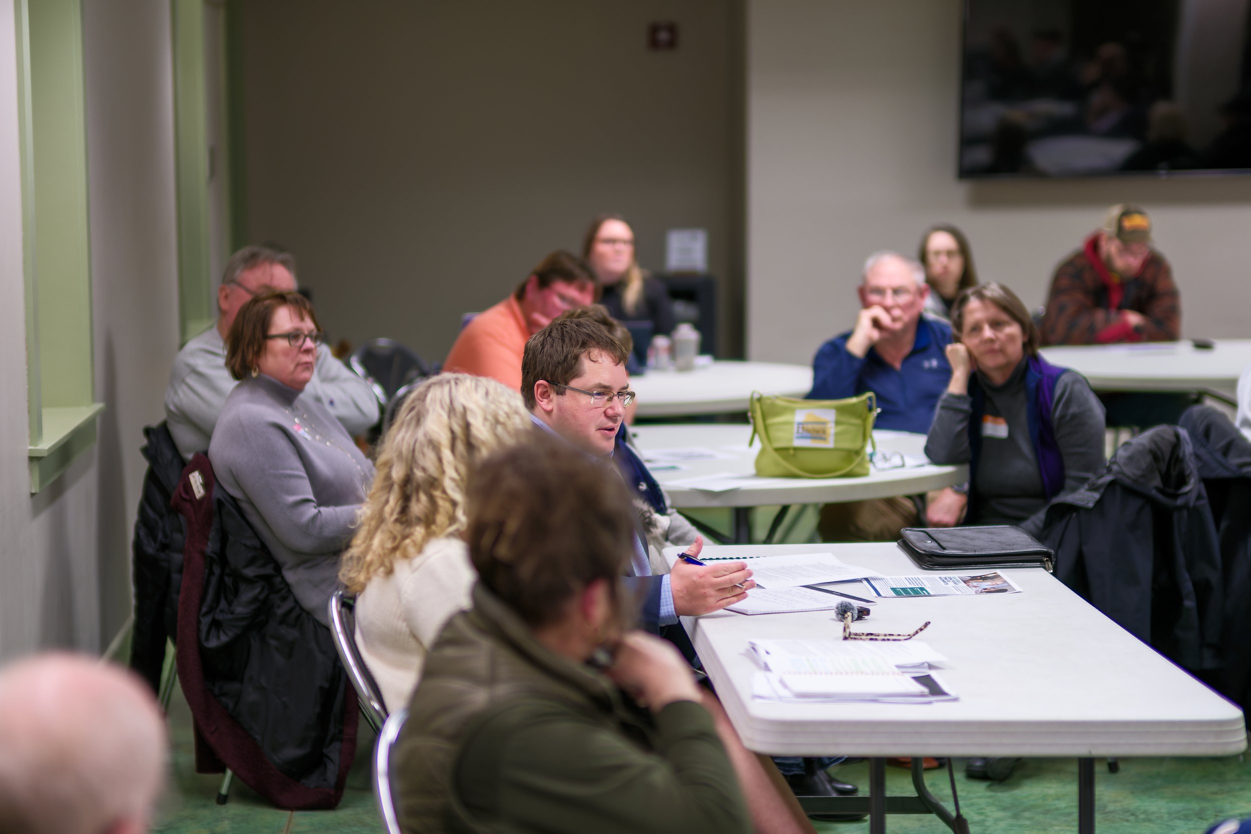 People attending a meeting or conference in a room with round tables, listening attentively, some taking notes, with a large screen on the wall in the background.