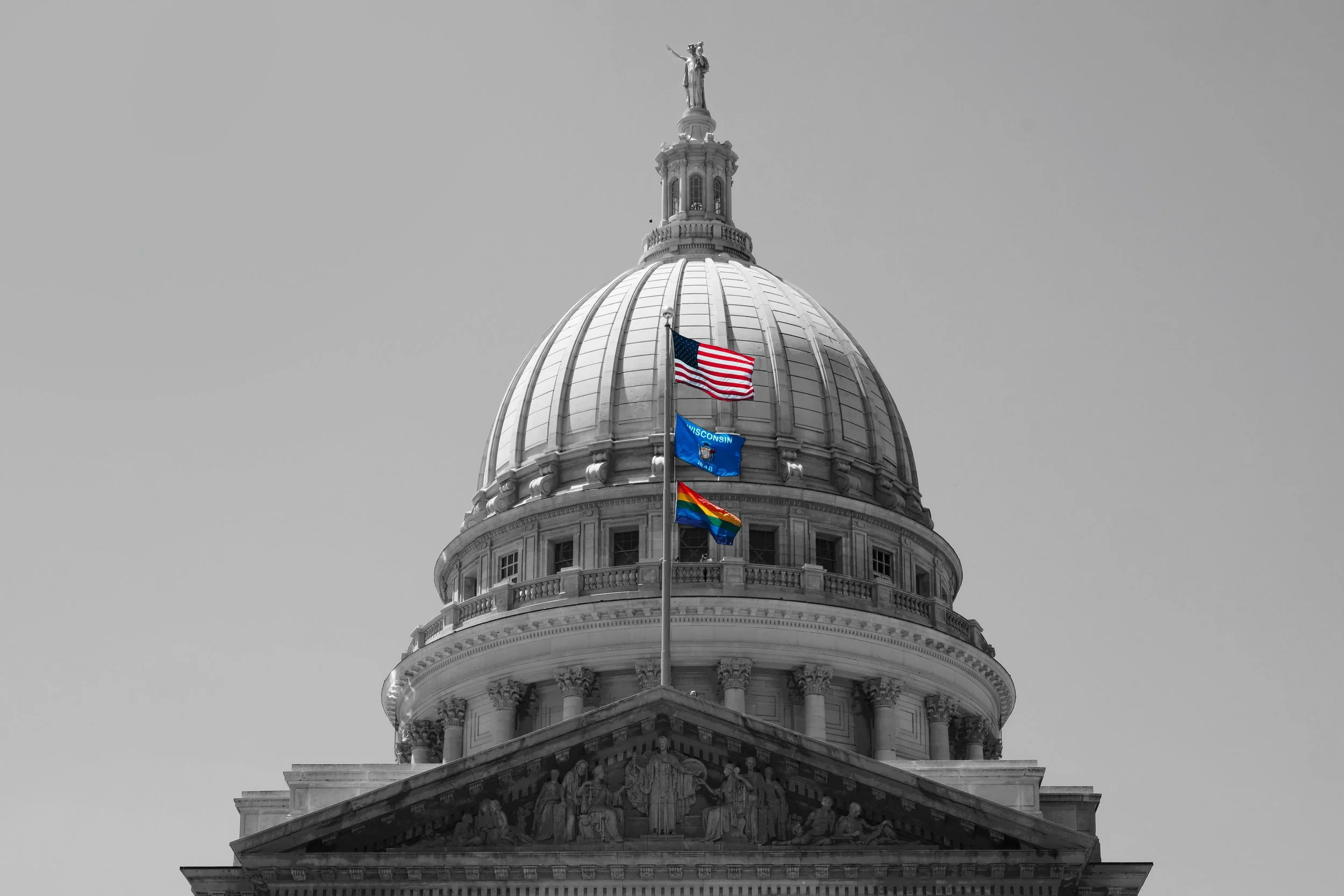 Close-up of the dome of the Wisconsin State Capitol with three flags flying in front: an American flag, the Wisconsin state flag, and a rainbow pride flag.