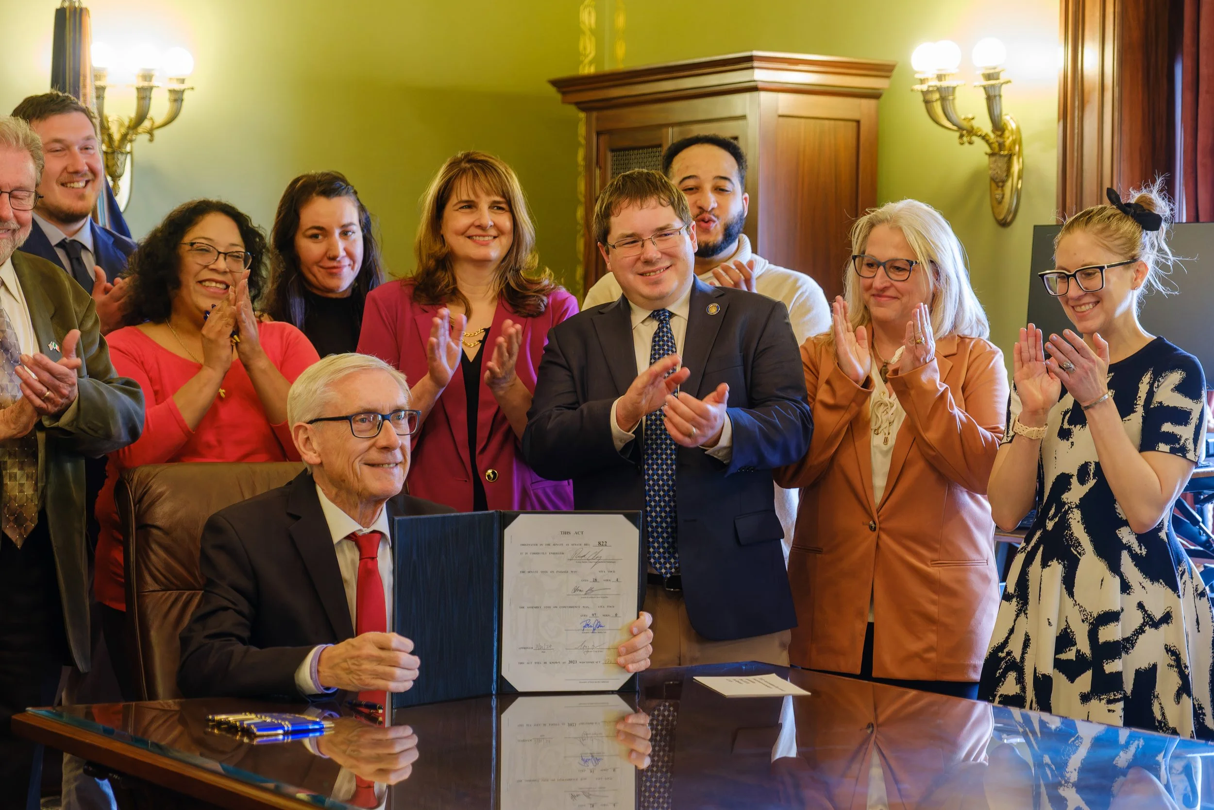 Group of people celebrating a signing ceremony, with Wisconsin Governor Tony Evers seated at a desk holding a signed bill, surrounded by applauding attendees in the Governor's office.